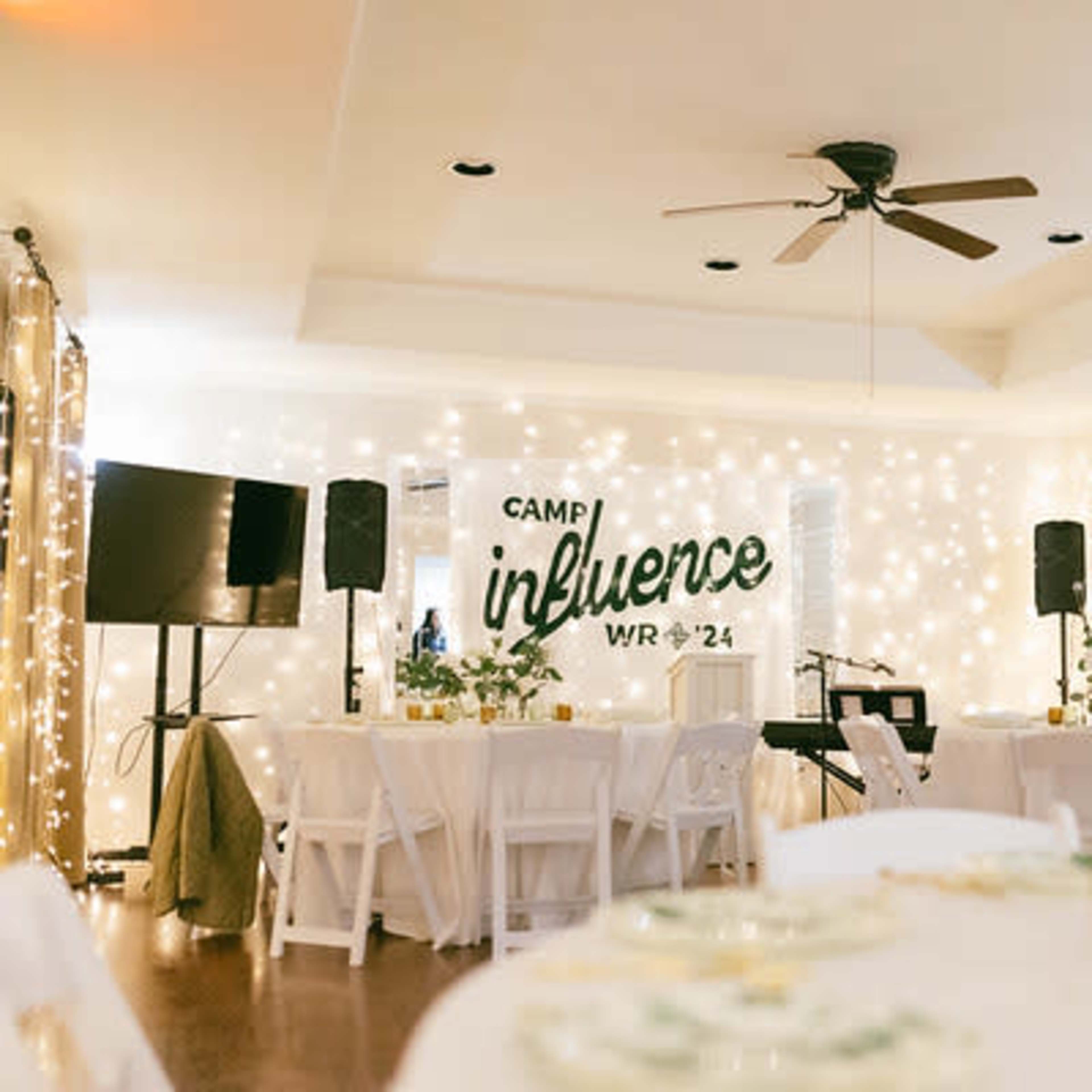 A decorated indoor event space featuring tables, string lights, and a backdrop with the words "Camp Influence WR 24."