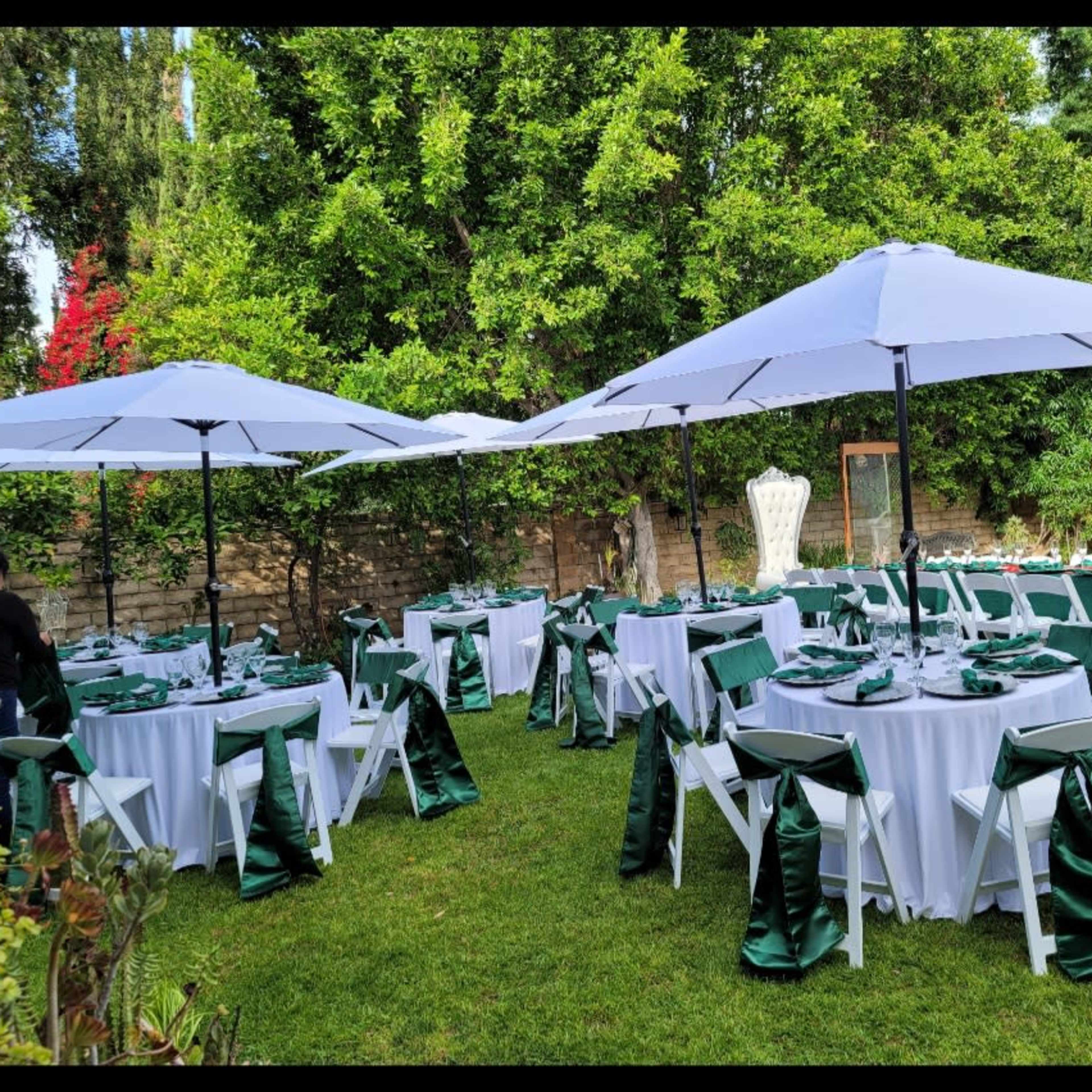 A neatly arranged outdoor event space features tables covered with white linens and decorated with green accents, shaded by large white umbrellas.