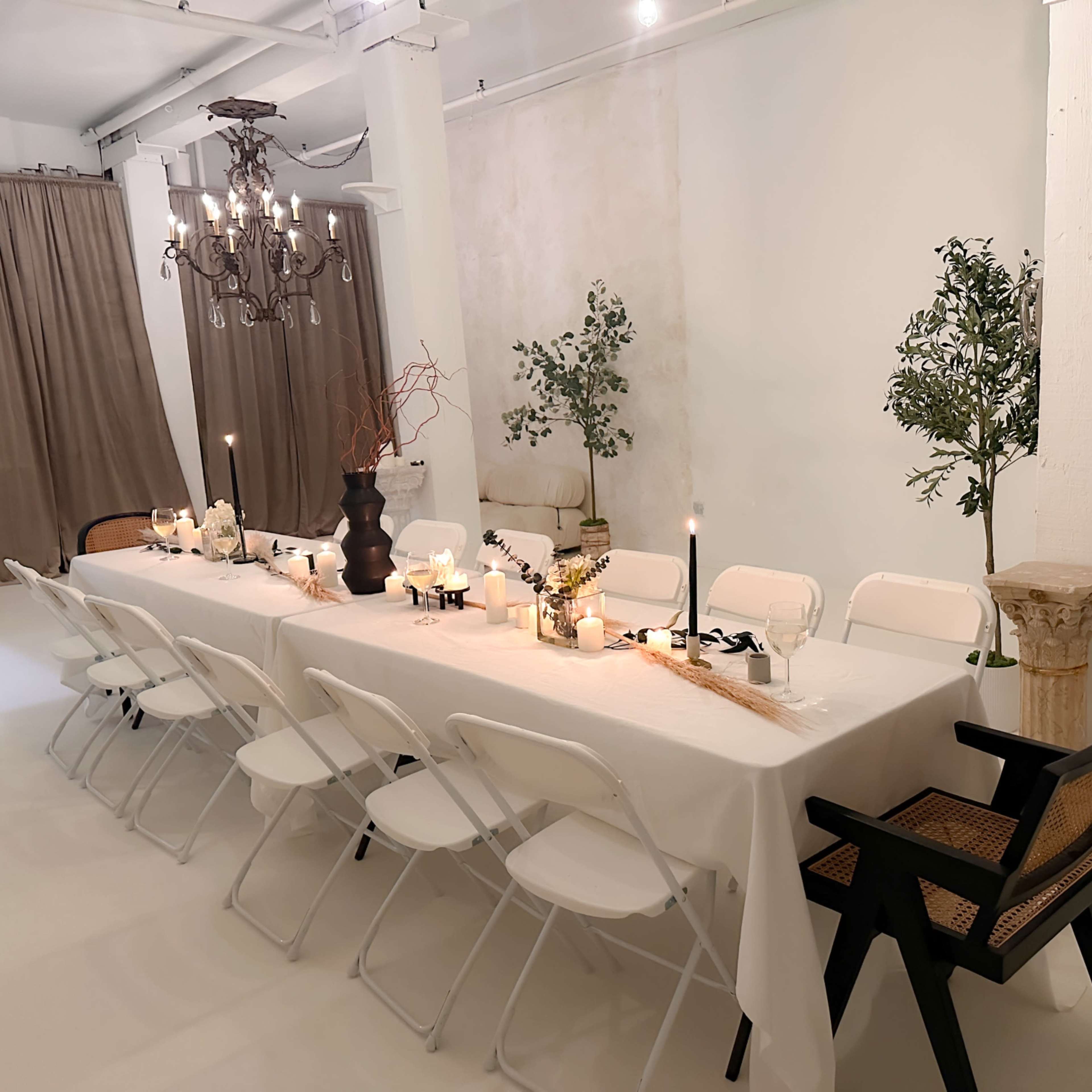A long white table set for a dinner with candles and decorative elements is positioned in a light-filled room with plants and curtains in the background.