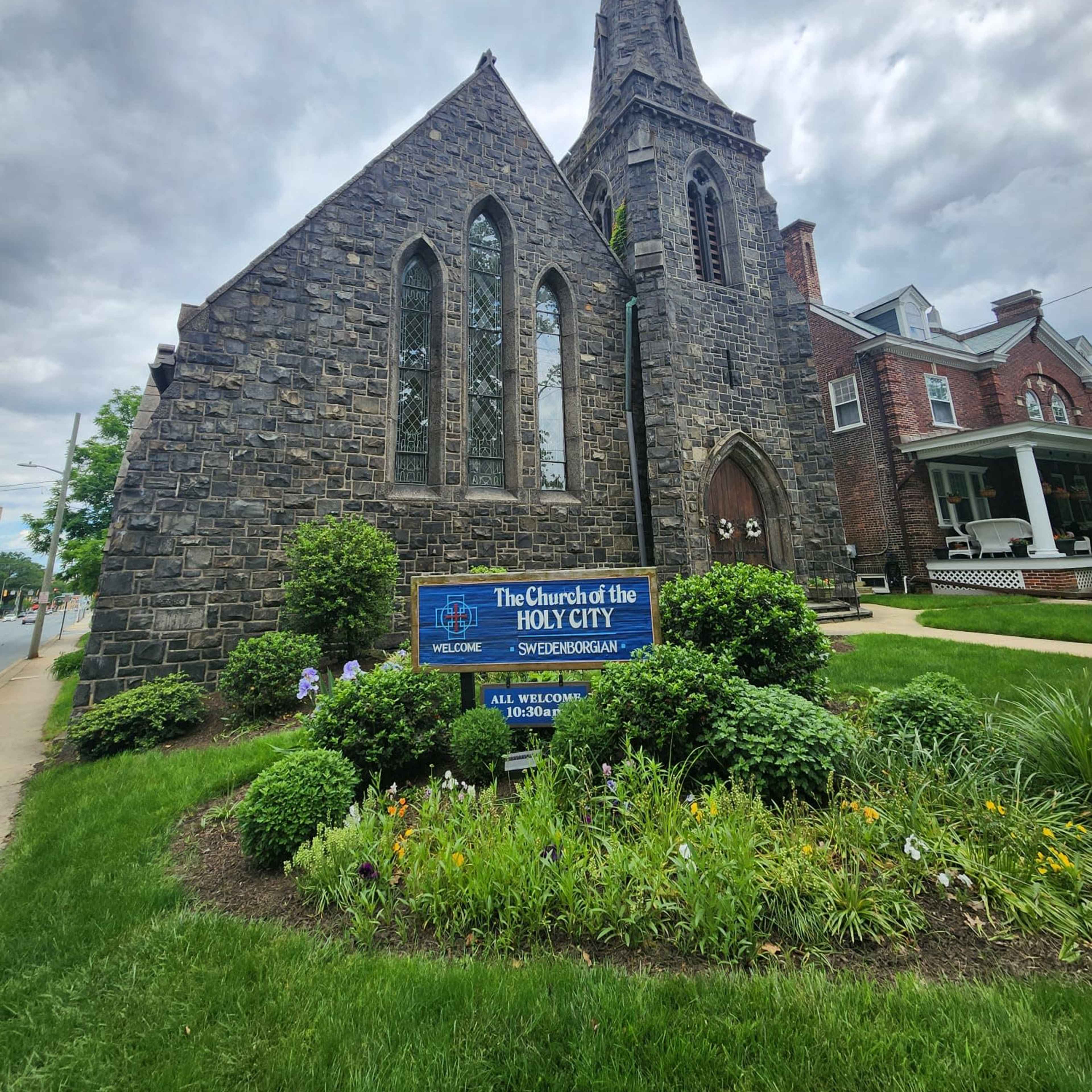 A stone church with a tall steeple, surrounded by greenery and a sign indicating it is the Church of the Holy City.