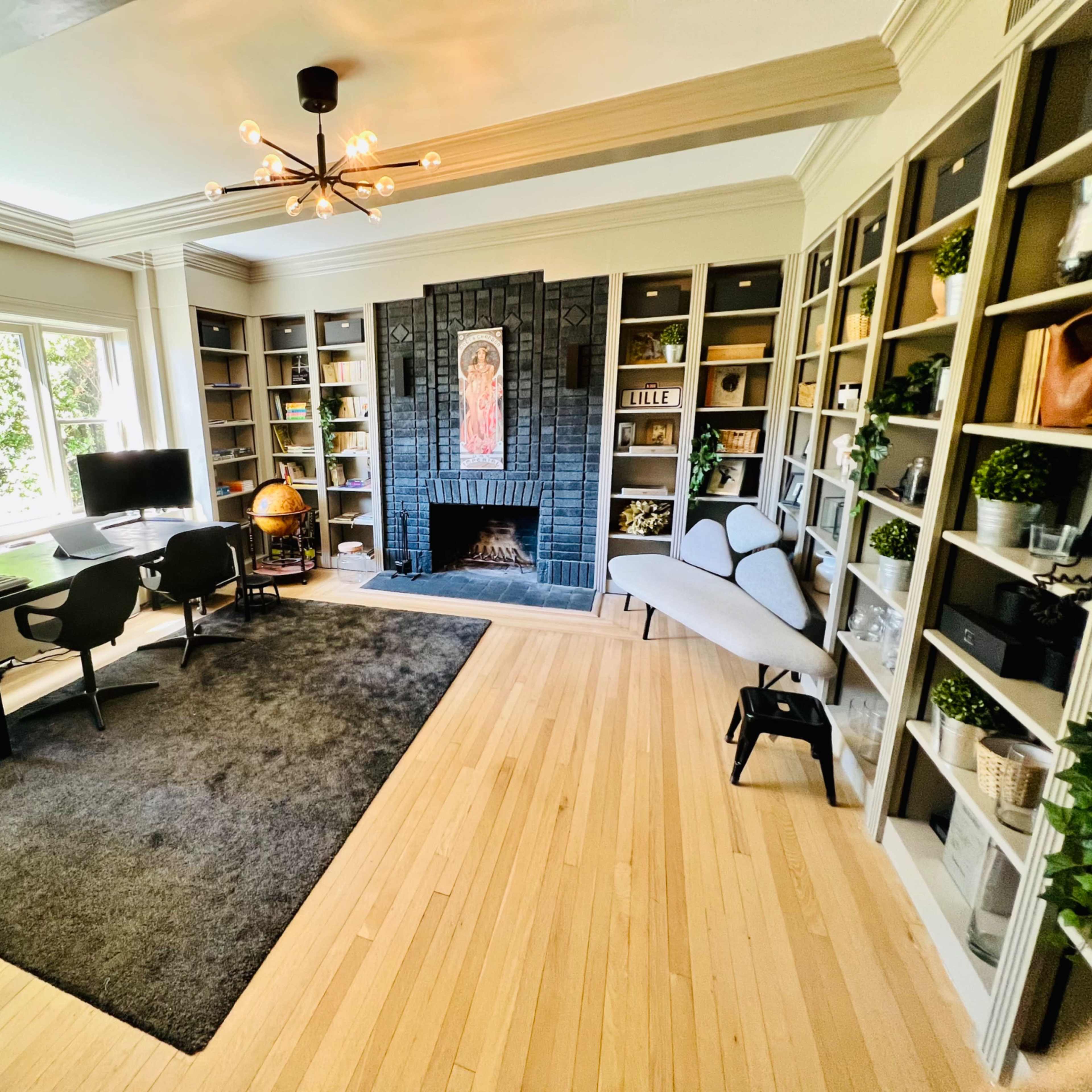A home office with a desk, two chairs, a large rug, bookshelves filled with plants and decor, and a fireplace against a black wall.