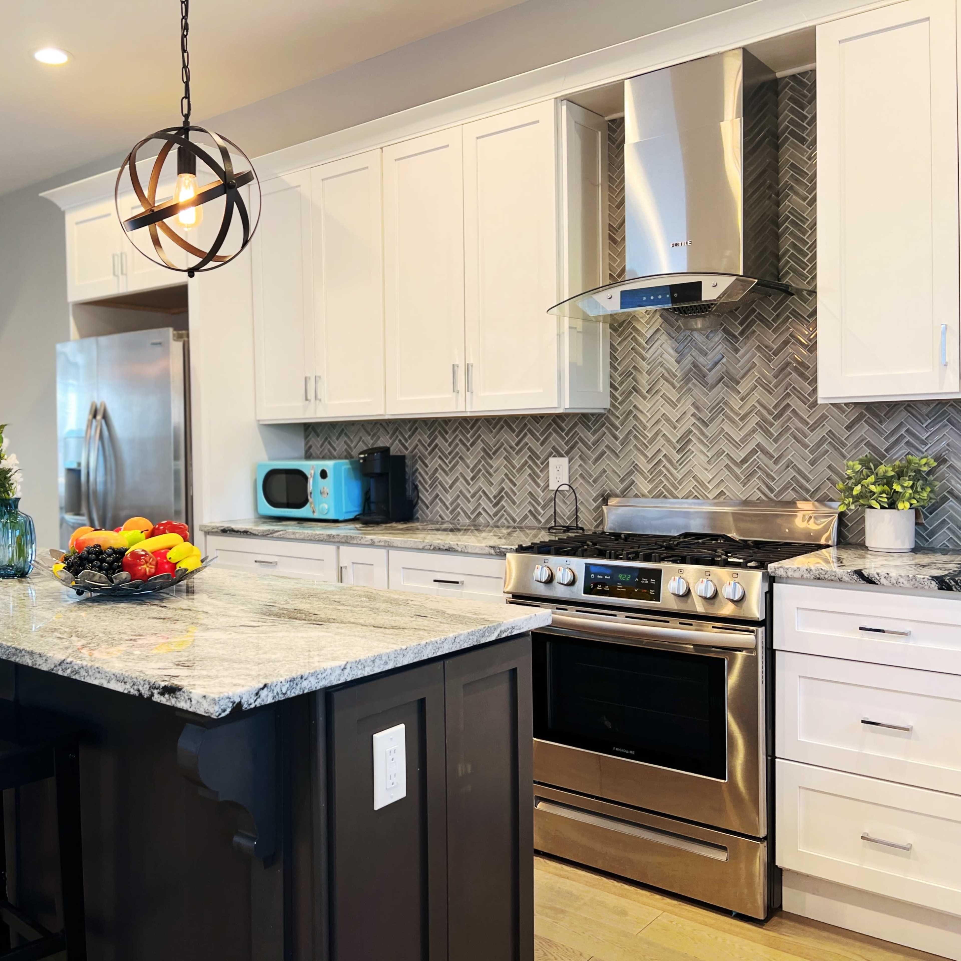 A modern kitchen with white cabinetry, stainless steel appliances, a gray and white herringbone backsplash, and a central island with bar stools and a fruit bowl.