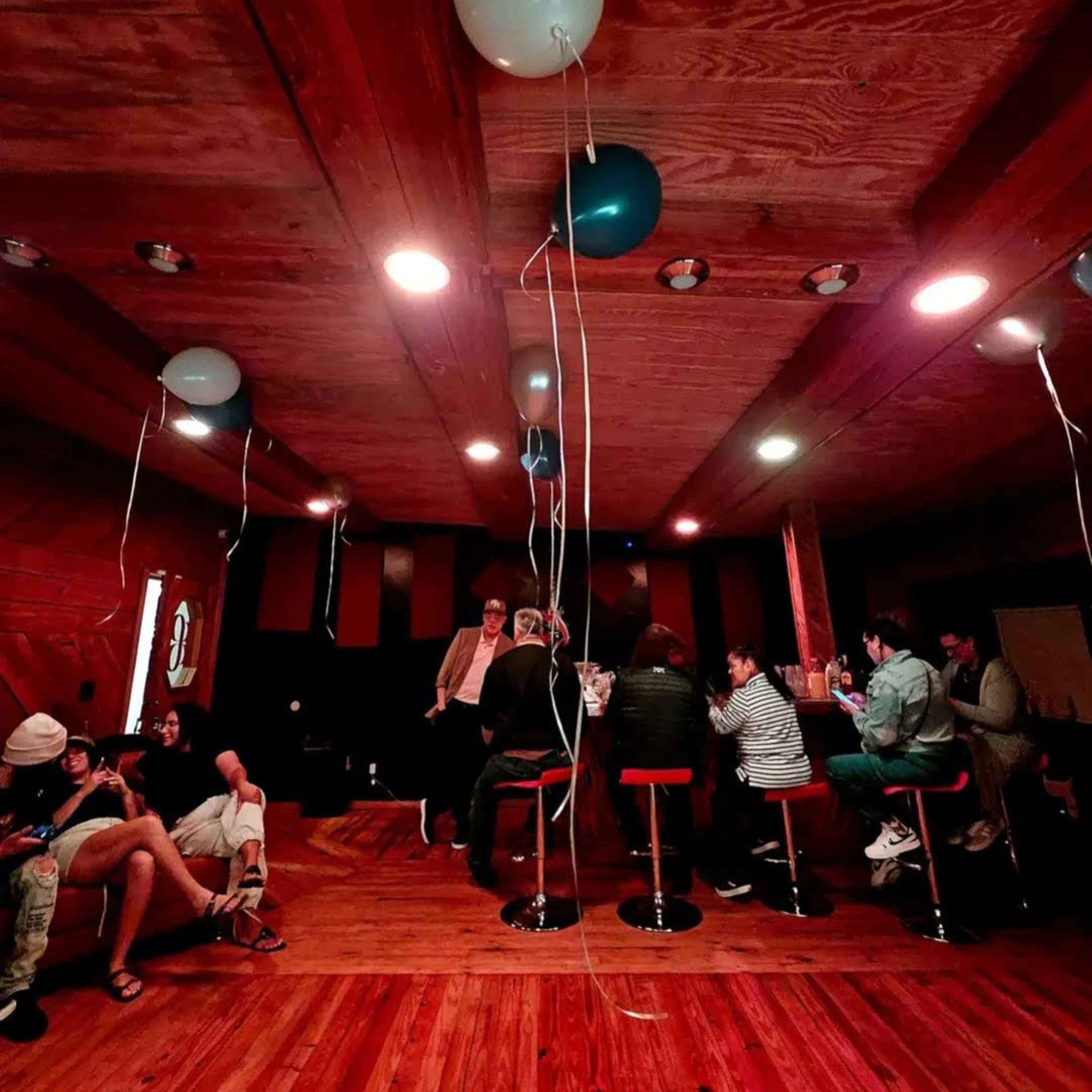 A wooden interior room with people seated at tables, decorated with balloons hanging from the ceiling.