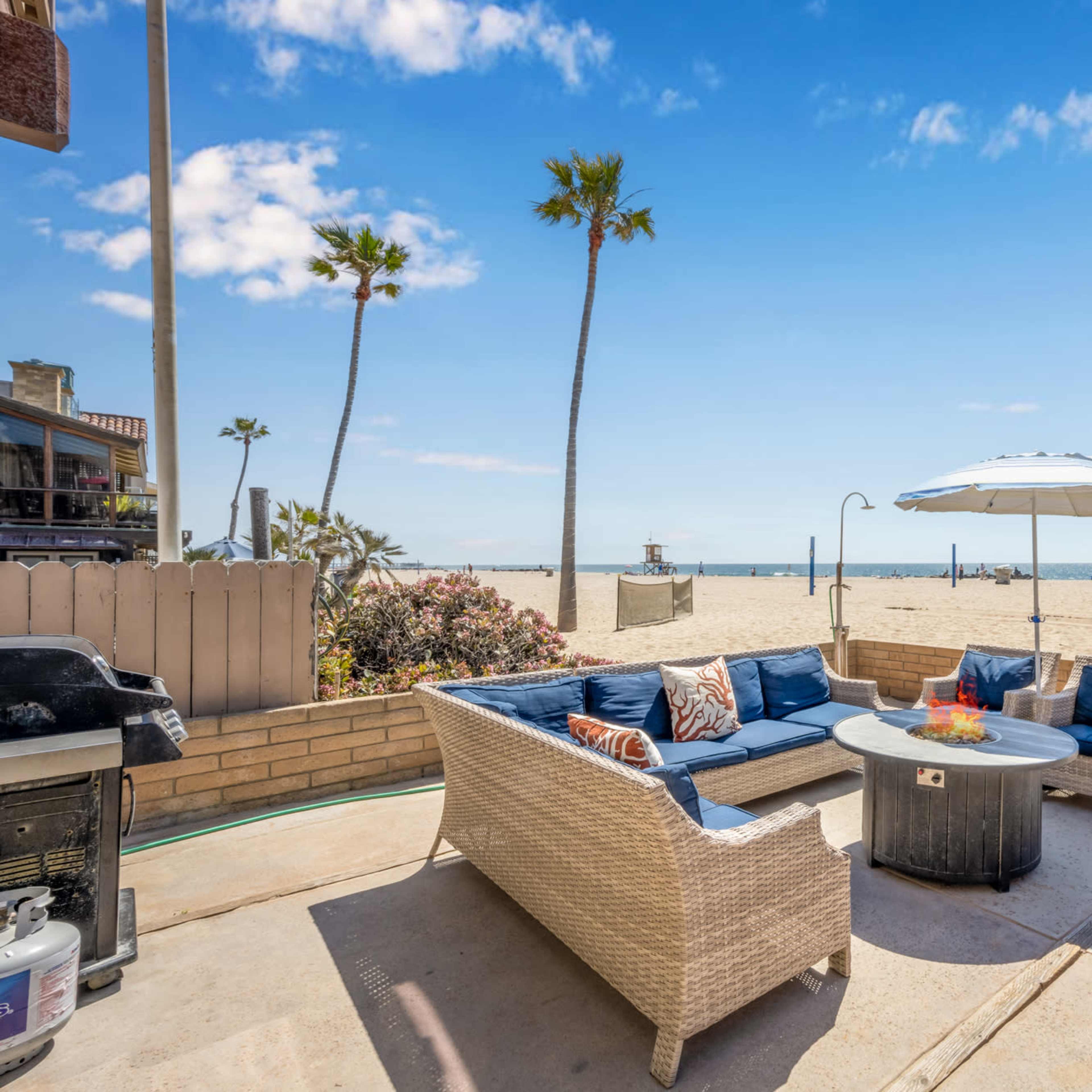 A patio area with a wicker sofa, a fire pit, and an umbrella overlooks a sandy beach and ocean, with palm trees in the background.