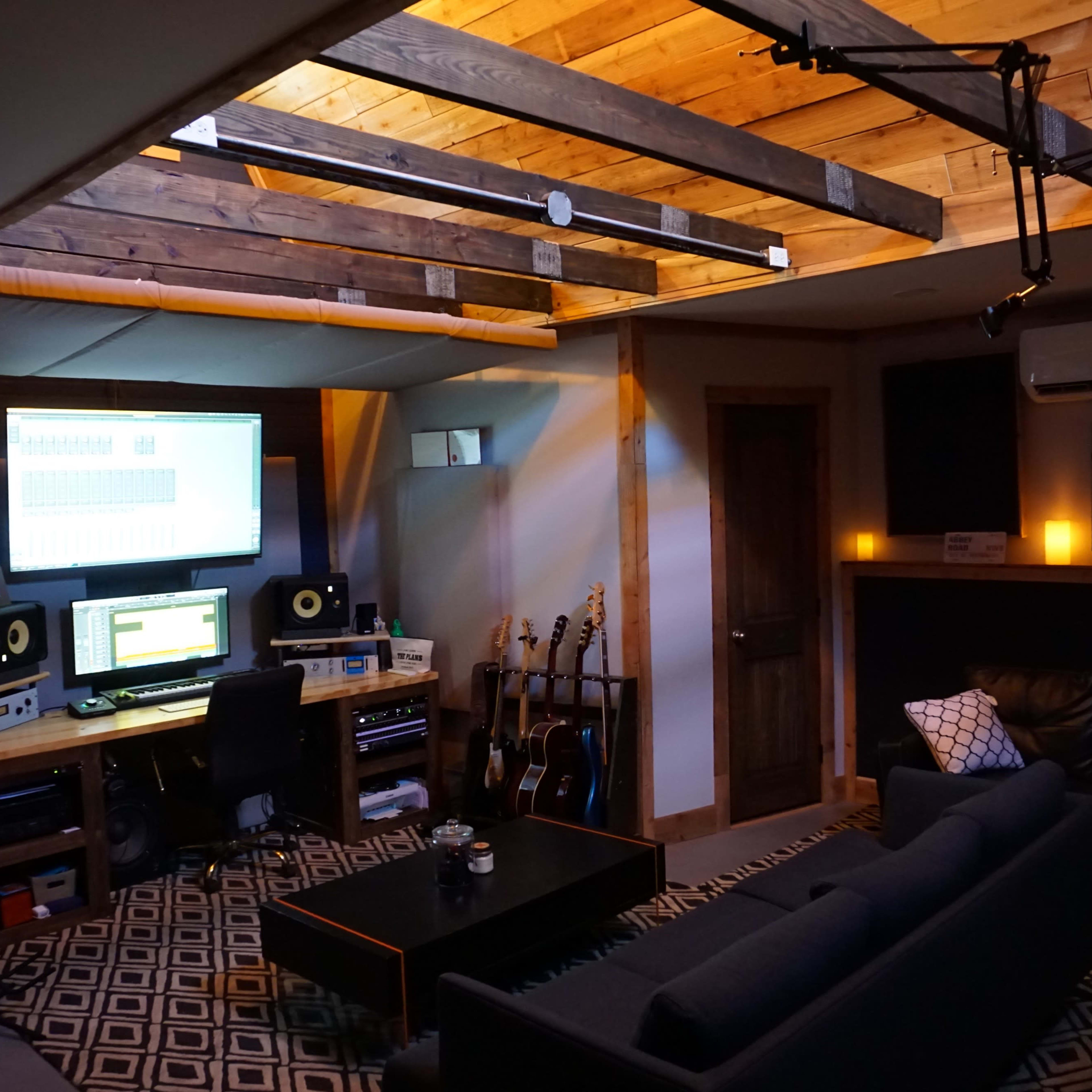 A cozy home recording studio featuring a desk with audio equipment, computer screens, guitars, and a comfortable seating area with a dark sofa.