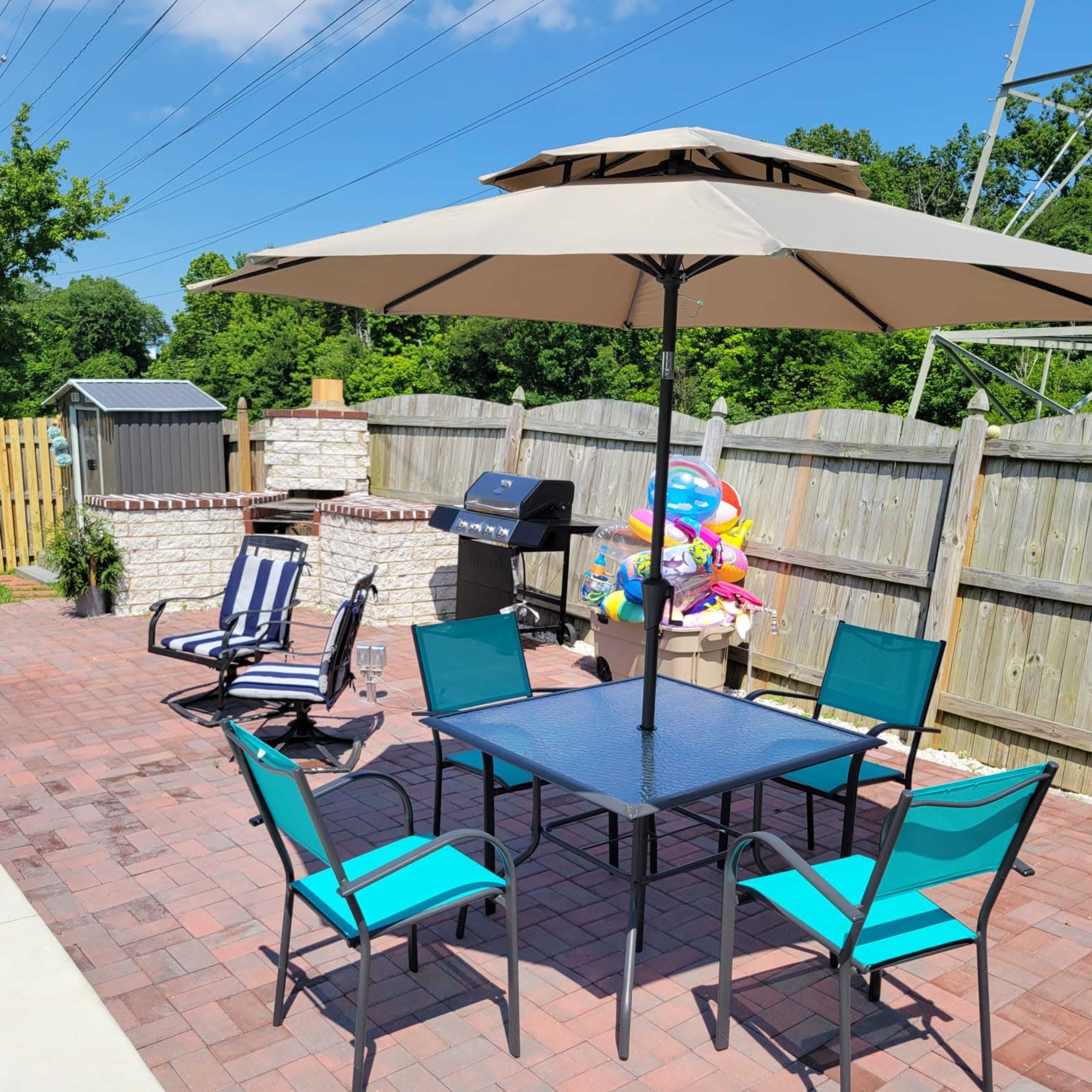 A patio area features a table with teal chairs, an umbrella, a grill, and a small storage shed, surrounded by a wooden fence and trees.