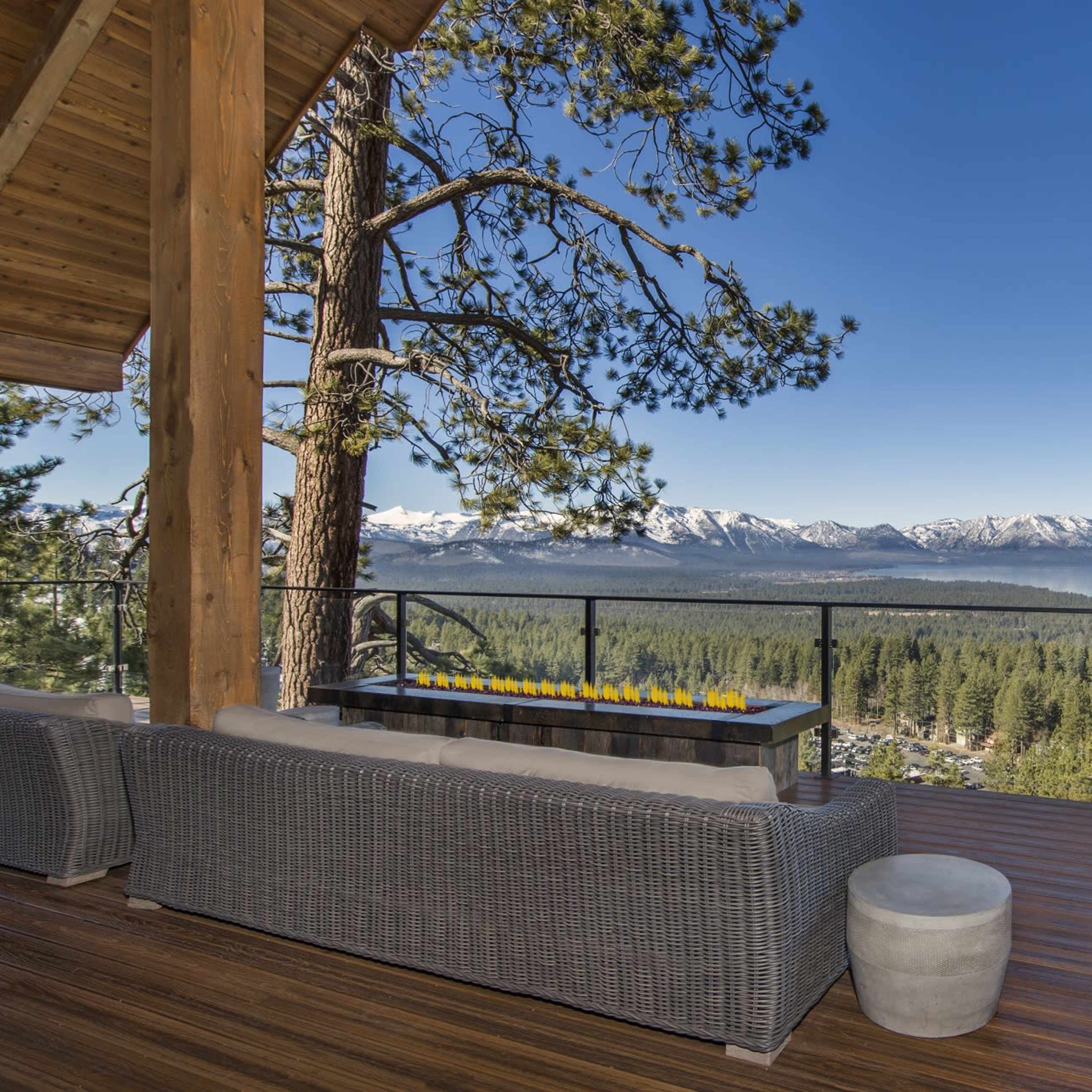 A spacious deck features comfortable seating and a fire table, overlooking a scenic view of mountains and a lake surrounded by trees.