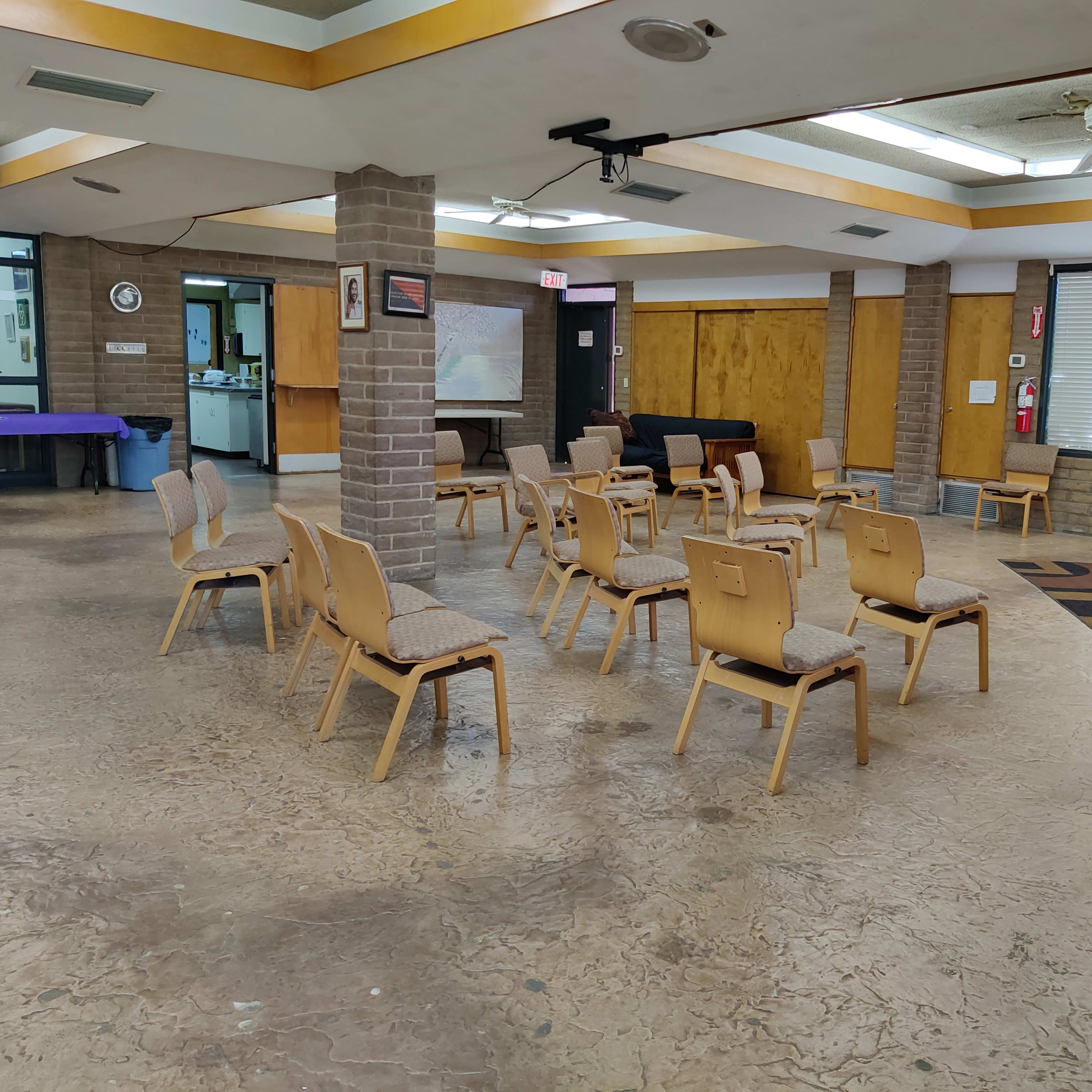 A room with several wooden chairs arranged in rows around a central space, featuring a podium with a cross and large windows along one side.