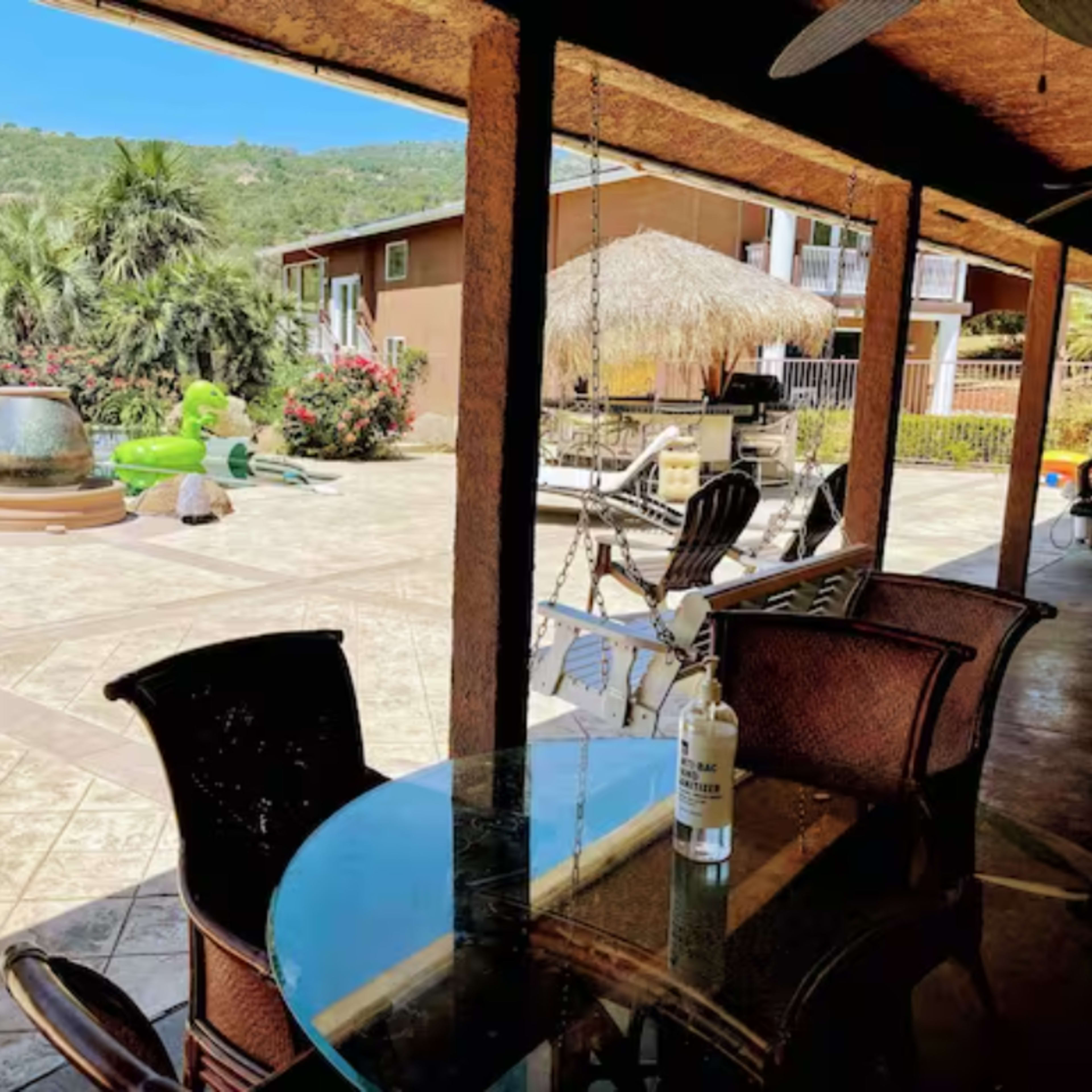 A patio with a glass table and chairs overlooking a swimming pool surrounded by tropical vegetation and a thatched roof structure.