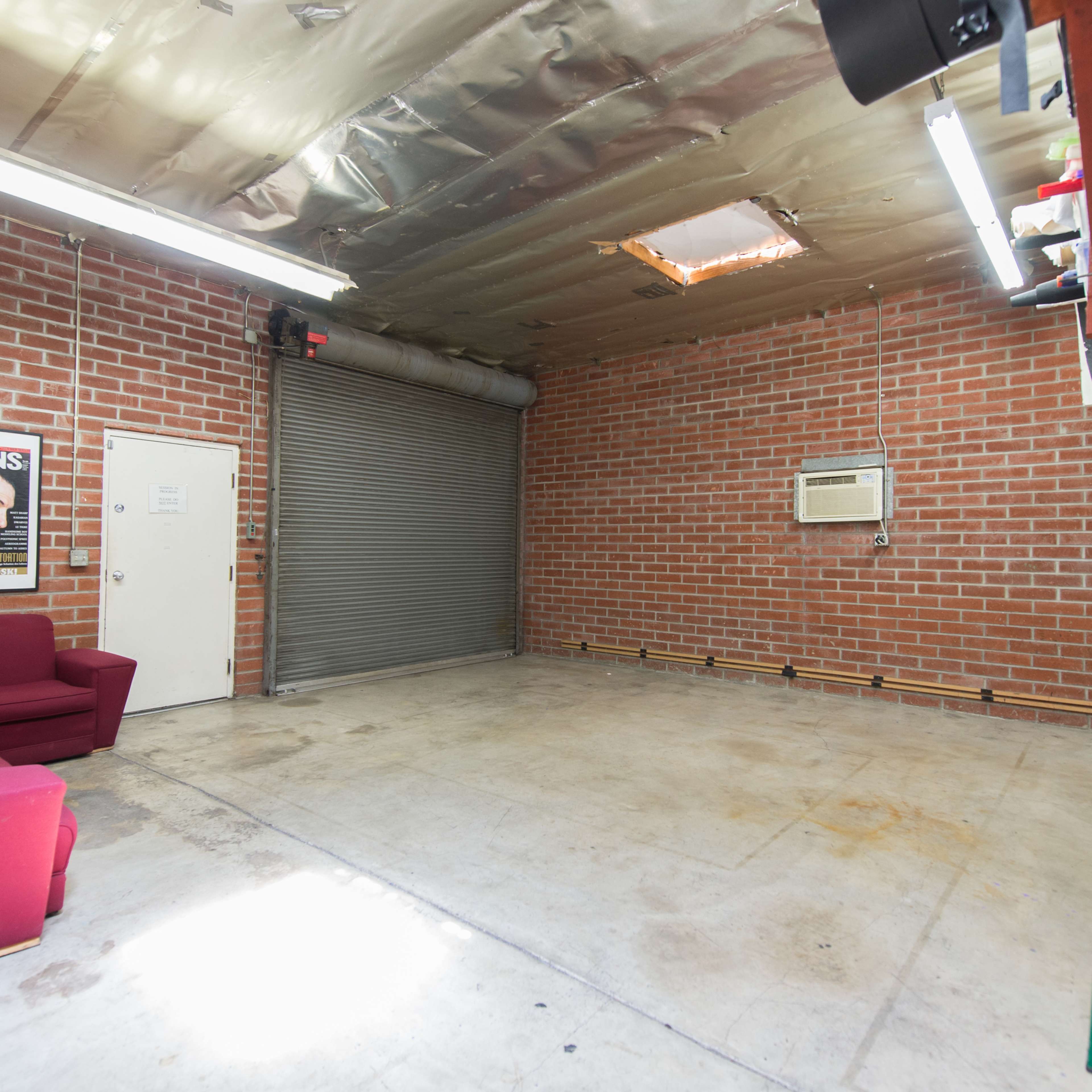 A spacious garage with exposed brick walls, a roll-up door, a wall-mounted air conditioner, and a maroon couch.