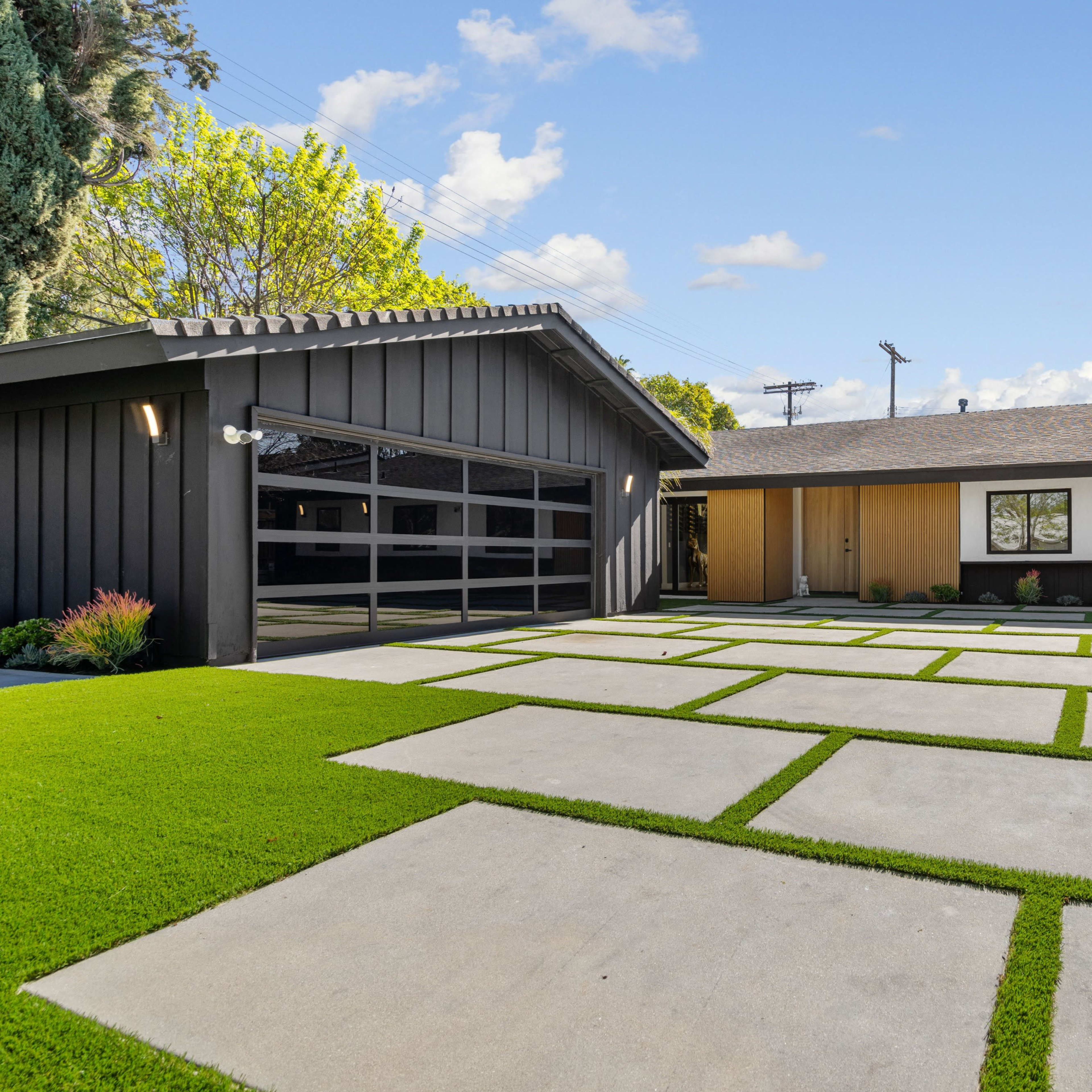 A modern house with a dark exterior features a concrete driveway surrounded by green grass and decorative plants.