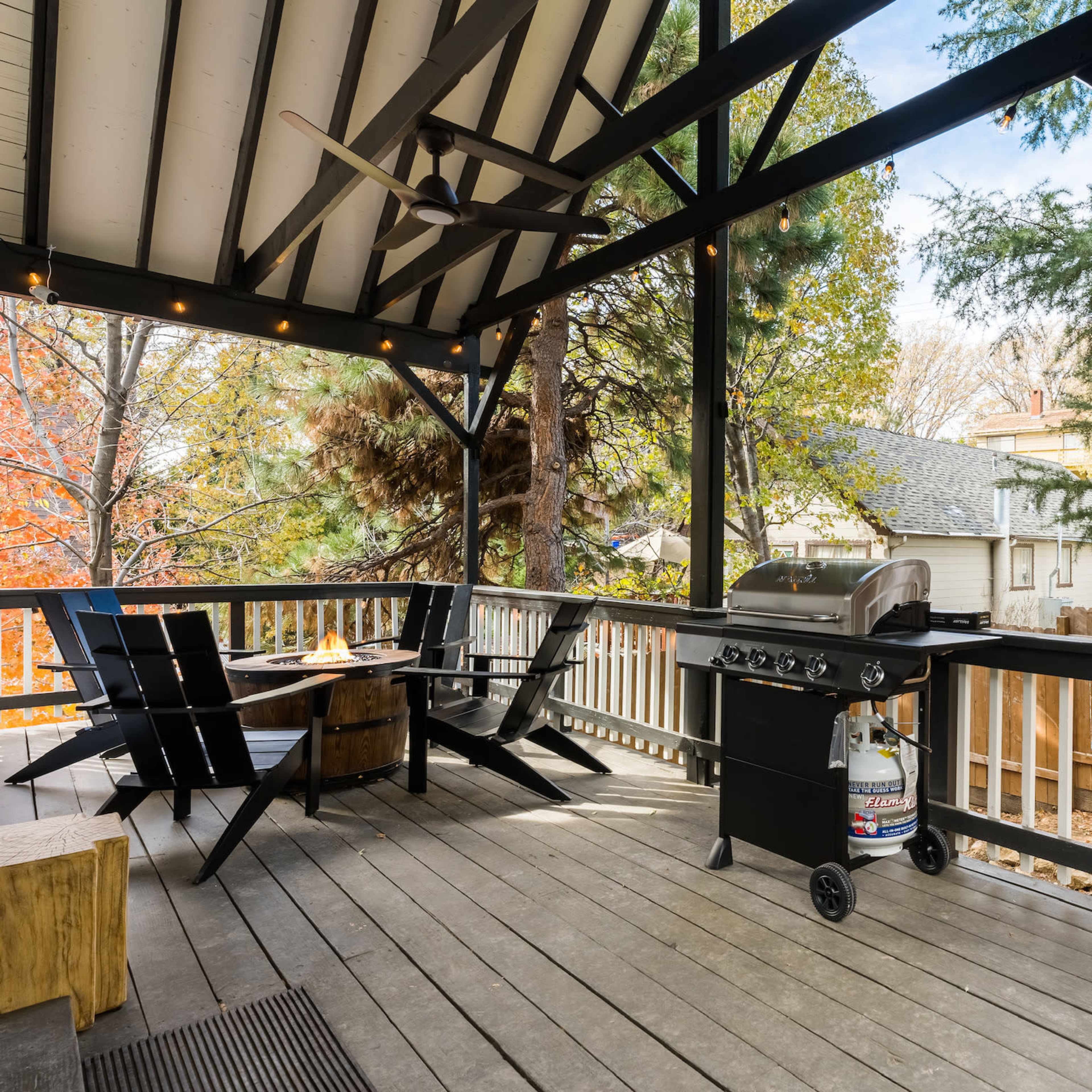 A covered deck featuring a gas grill, seating area with black chairs, and surrounding trees.
