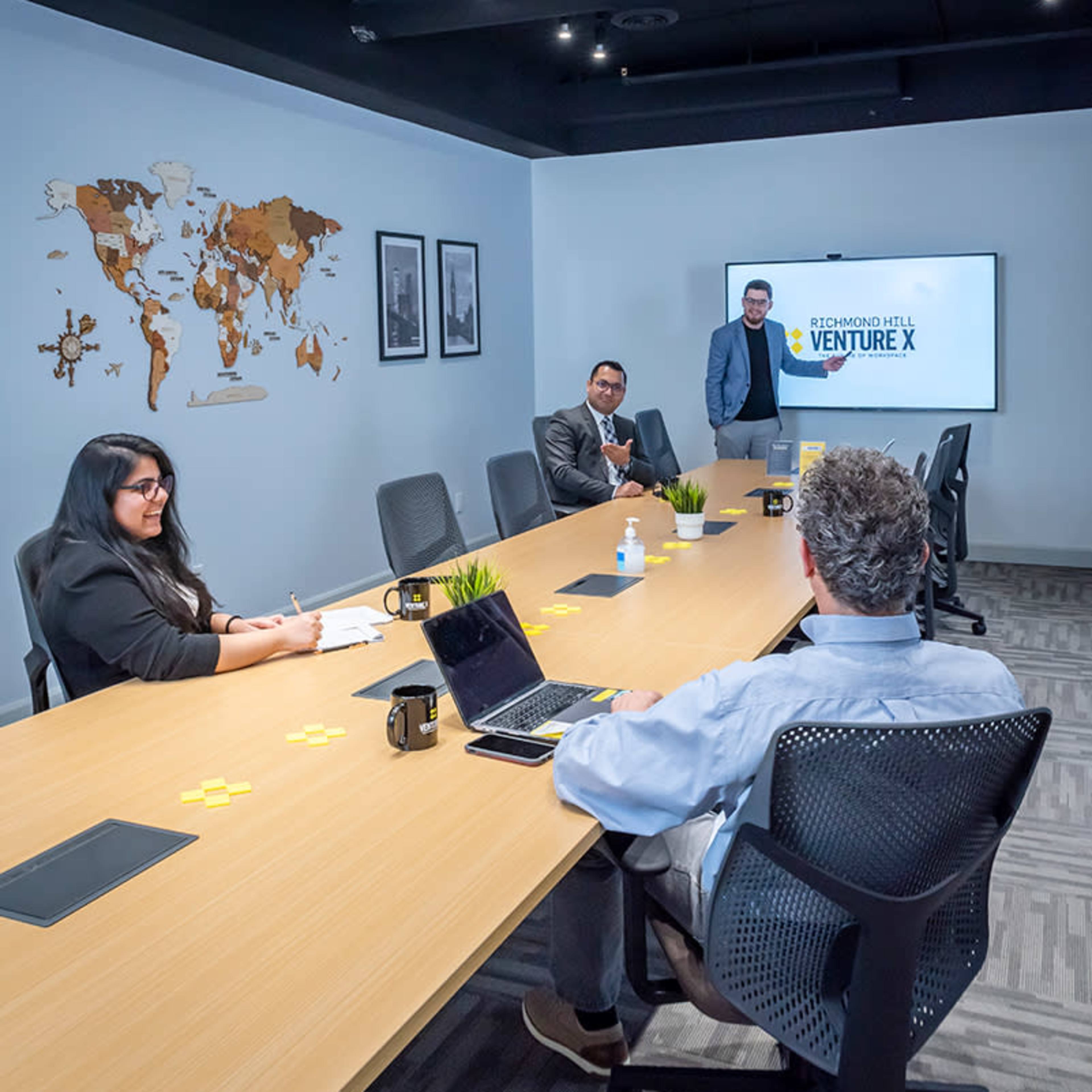A business meeting is taking place in a modern conference room with a presentation on a screen and a world map on the wall.