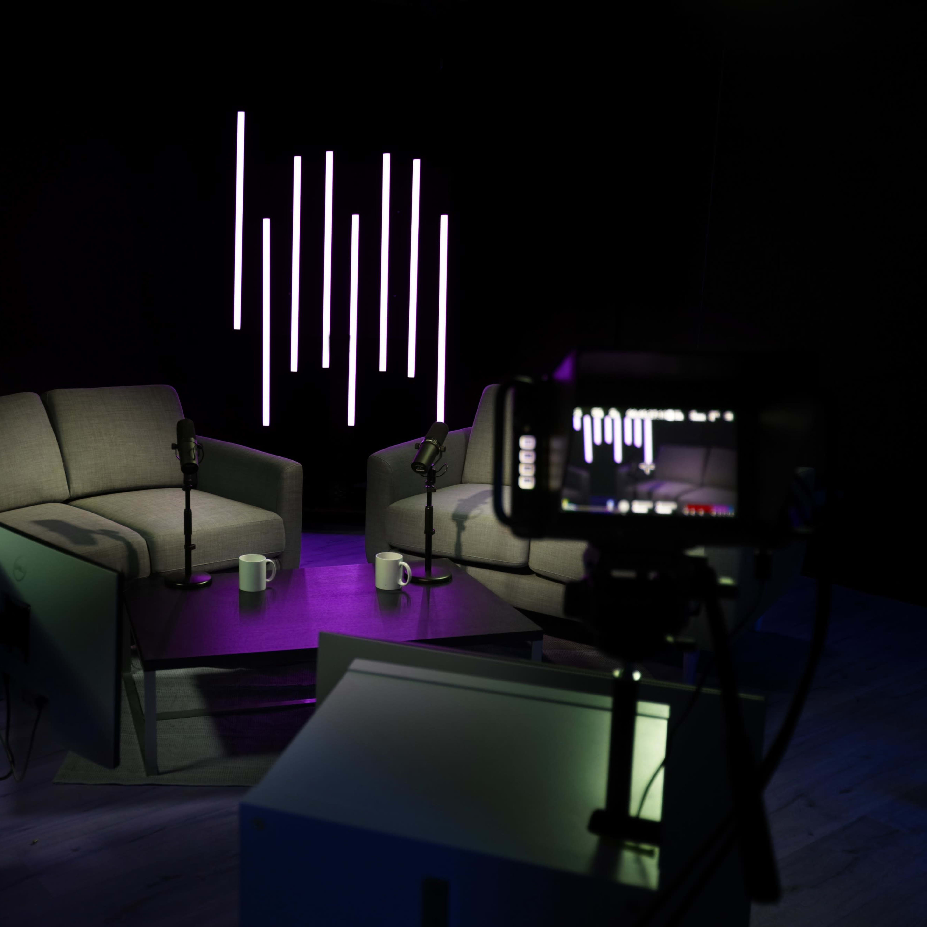 A dimly lit studio features two gray couches facing each other, with microphones and coffee mugs on a small table, alongside a camera and illuminated vertical light strips in the background.