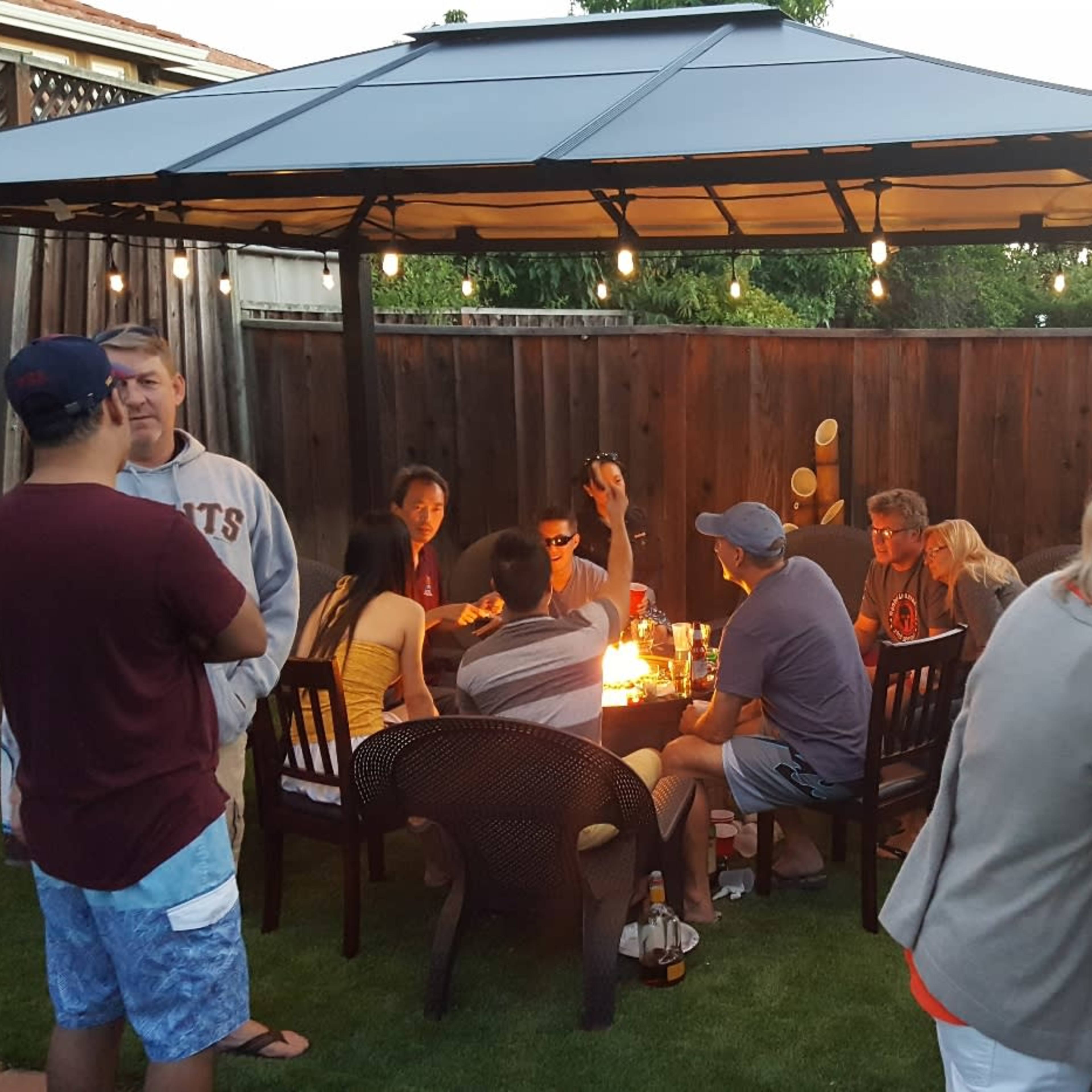 A group of people gathers around a table under a gazebo in a backyard, sharing food and conversation as evening sets in.