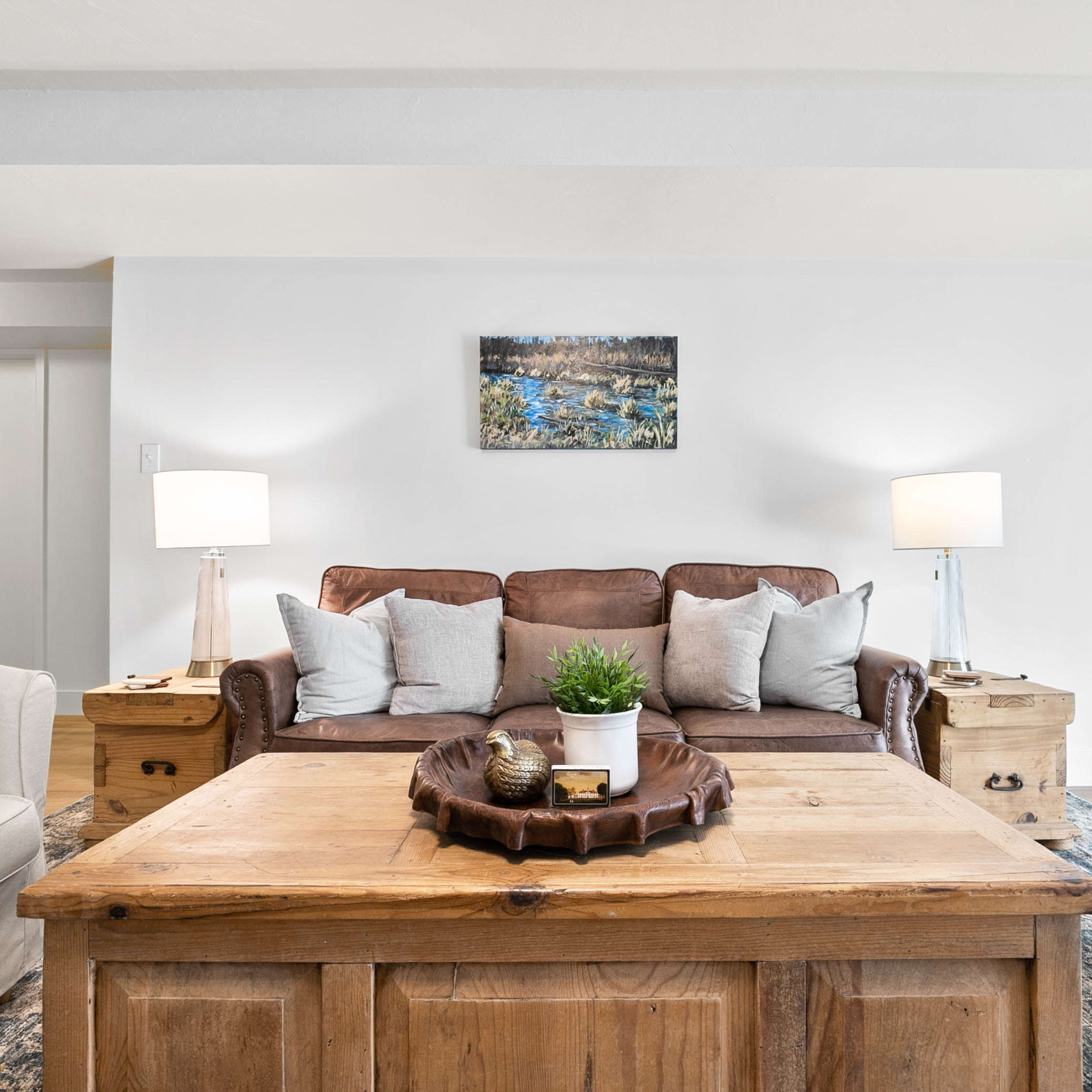 A living room features a brown leather sofa with decorative pillows, flanked by two light-colored chairs and a wooden coffee table, all set against a neutral wall and complemented by lamps and a piece of artwork.