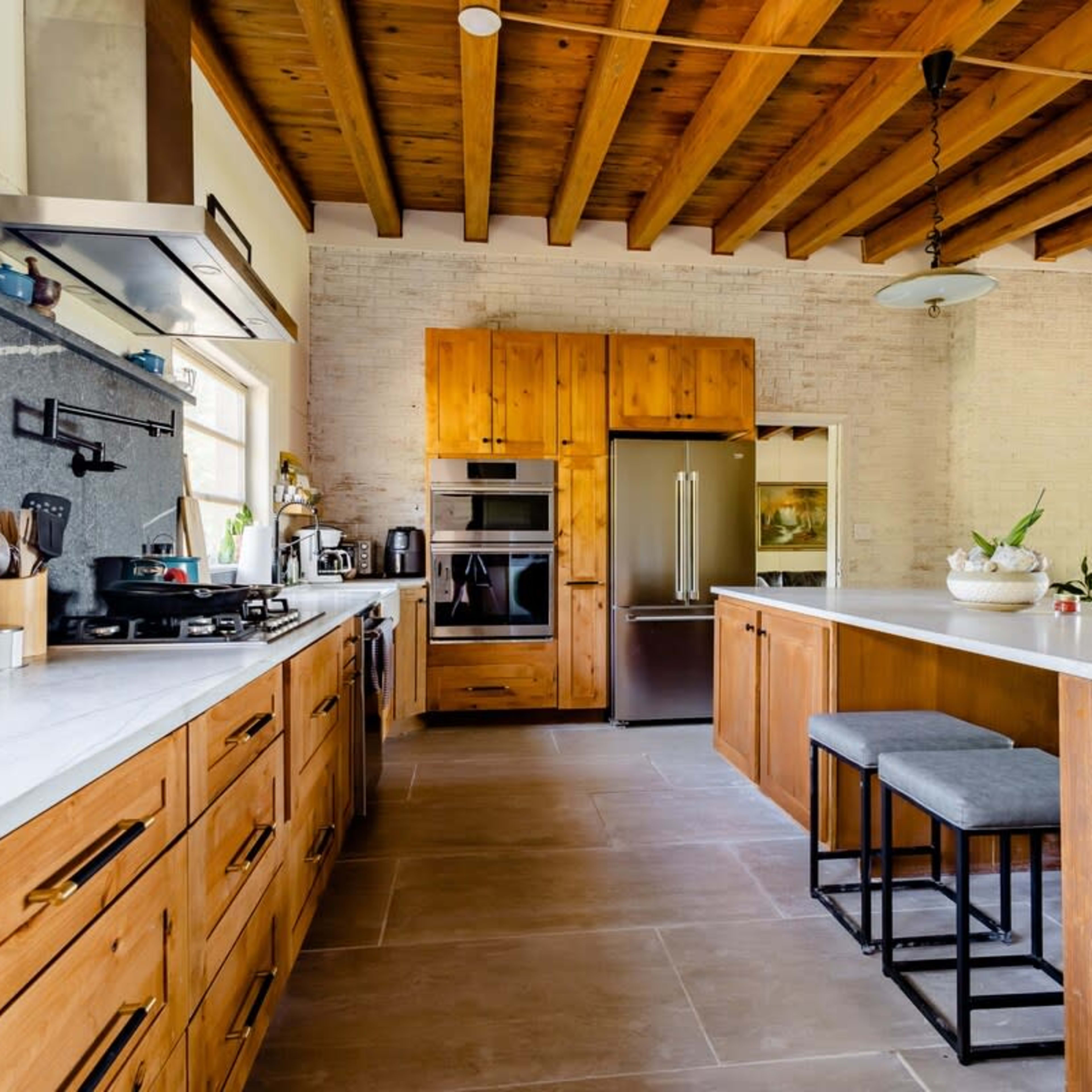 A modern kitchen features wooden cabinetry, a large island with bar stools, stainless steel appliances, and exposed wooden beams on the ceiling.