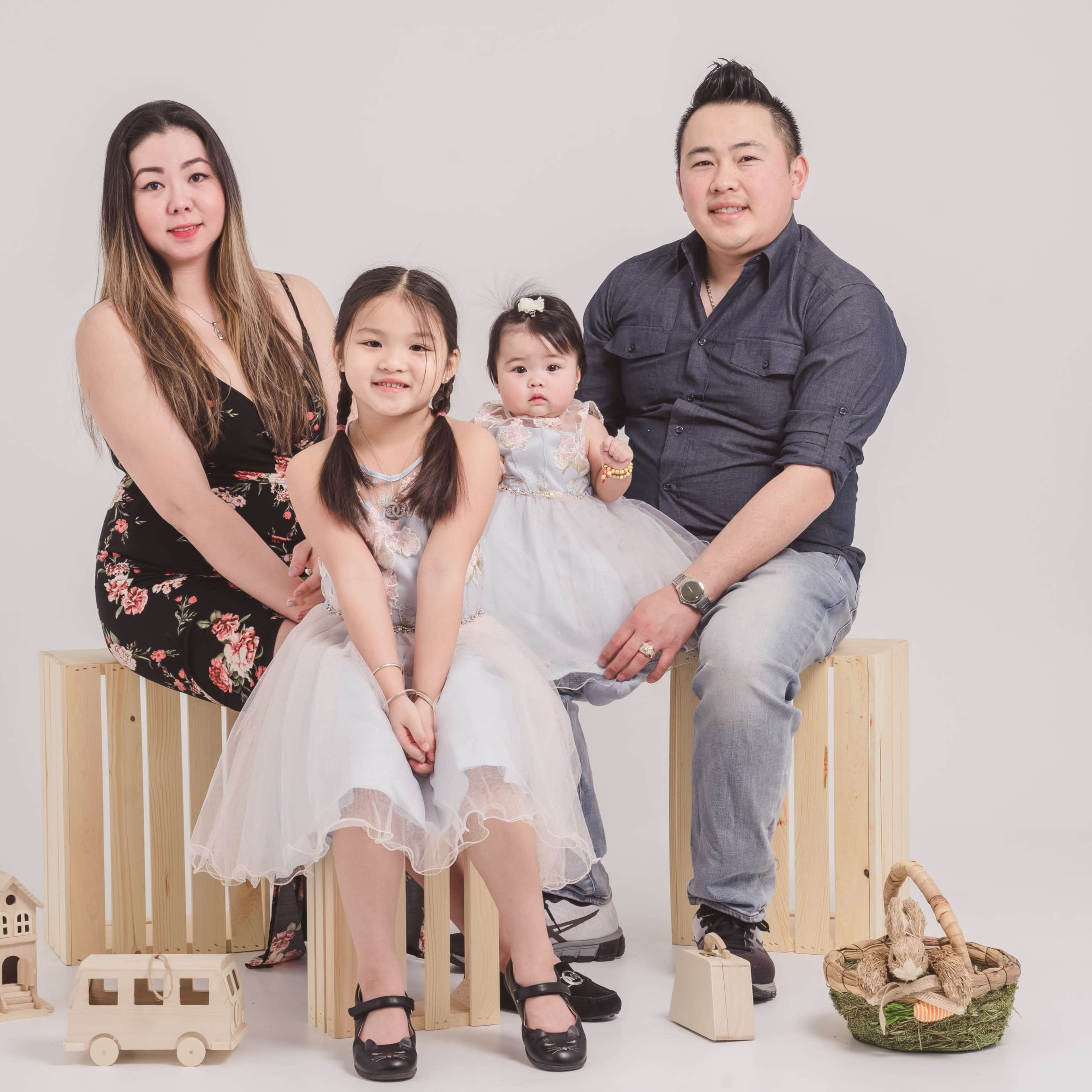 A family of four, consisting of two adults and two young girls, poses together on wooden crates with various wooden toys displayed around them.