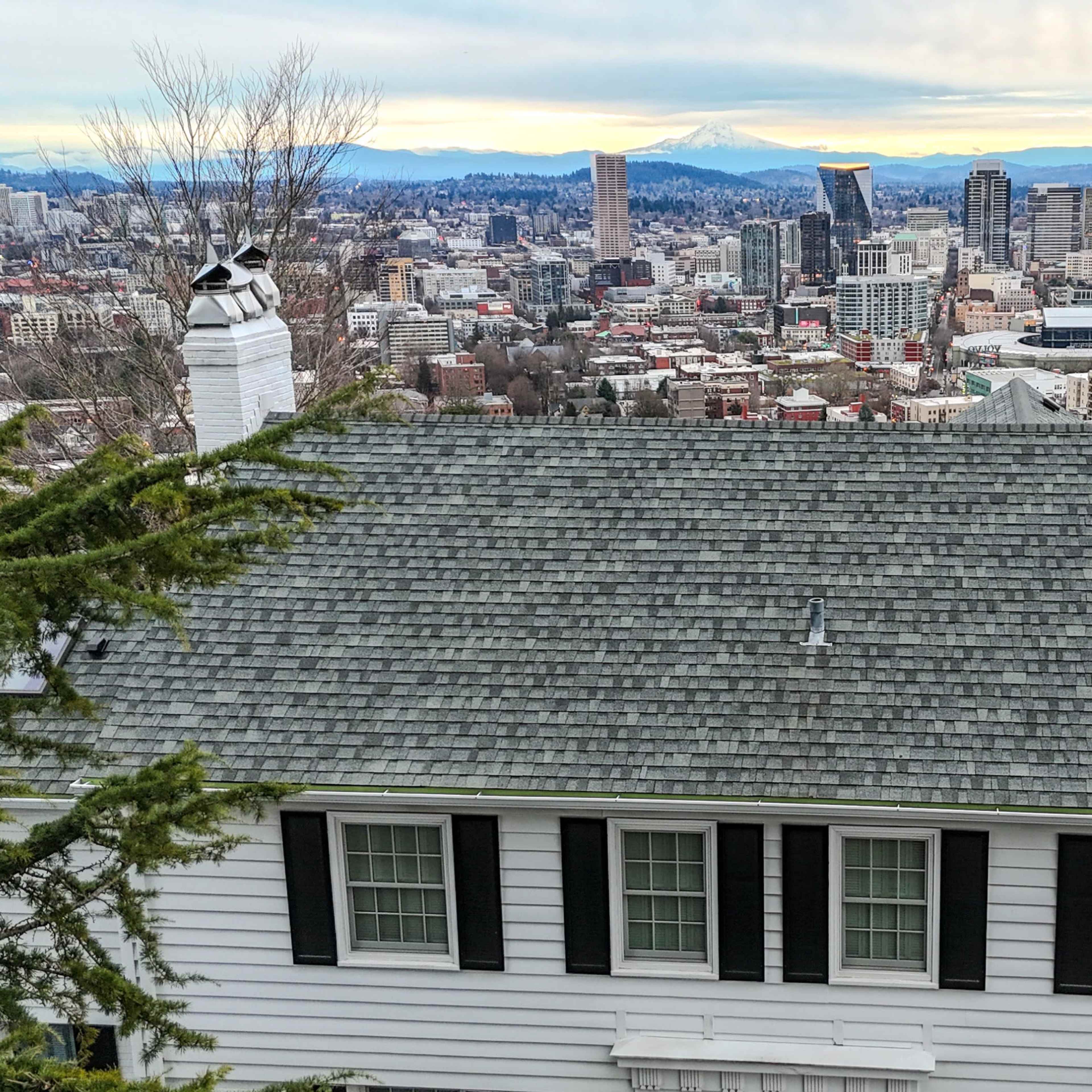 A white, two-story house with a gray shingle roof overlooks a city skyline featuring tall buildings and distant mountains.