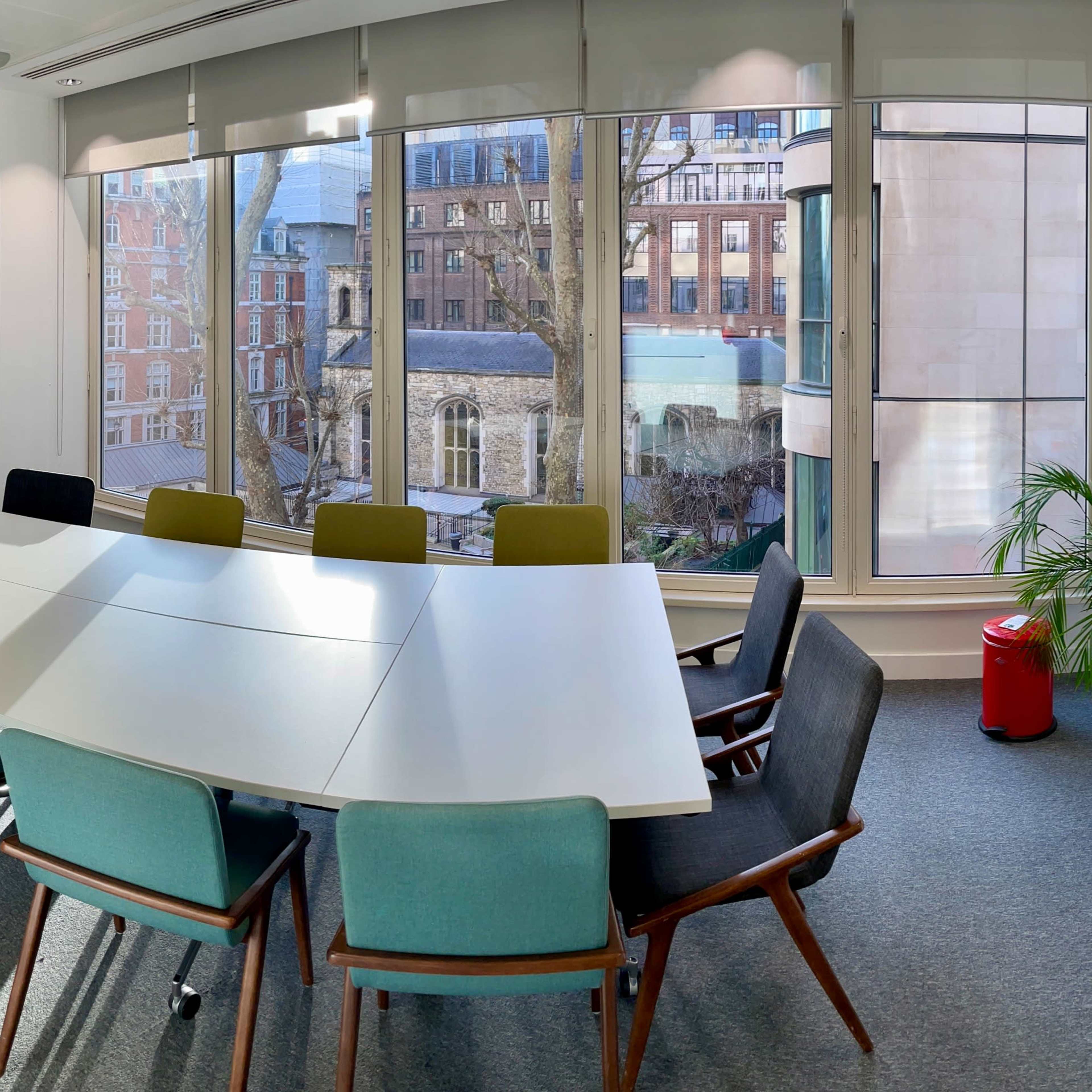 A modern conference room features a large white table surrounded by colorful chairs, with large windows overlooking an urban courtyard.