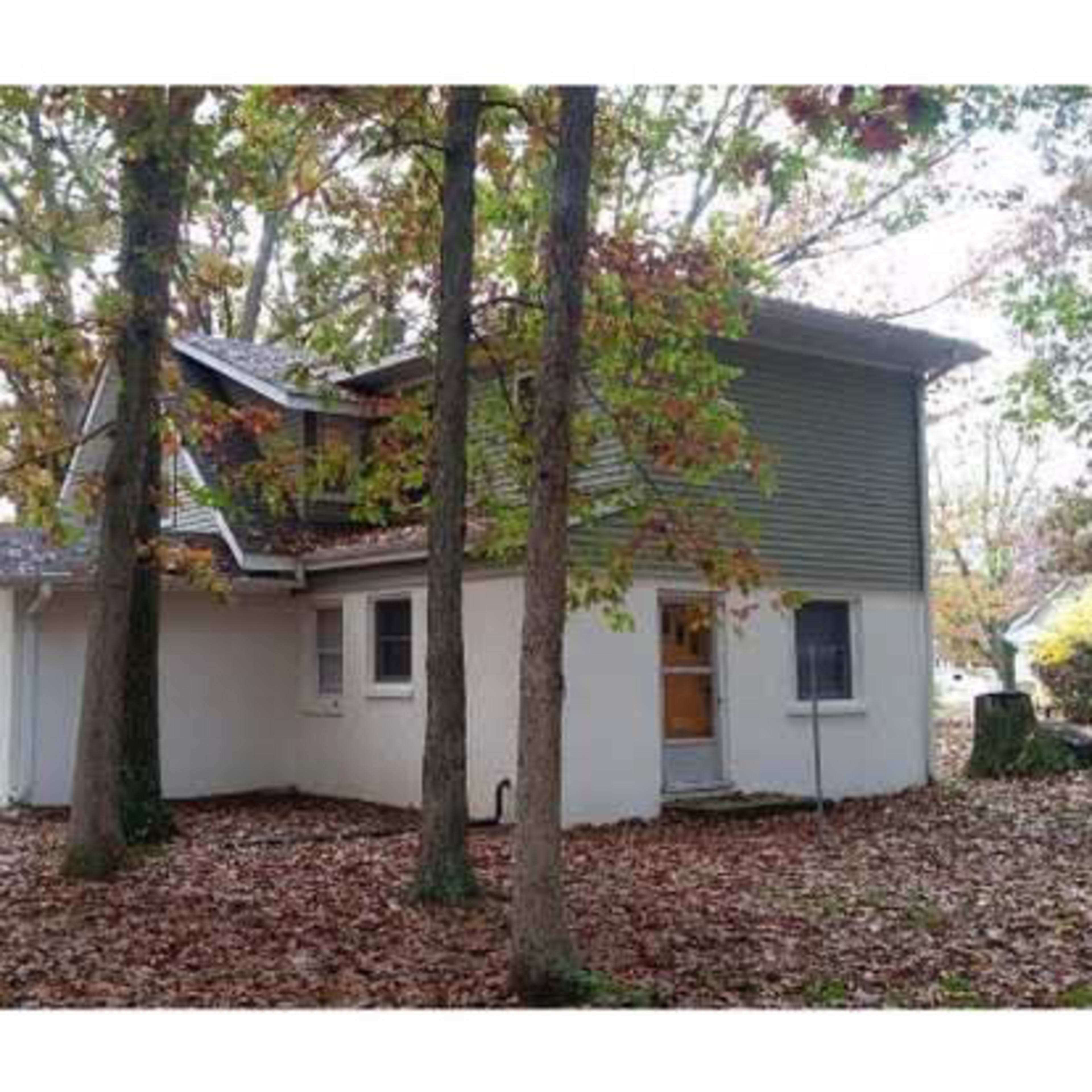 A two-story house with a green and gray exterior is surrounded by trees and fallen leaves in a wooded area.
