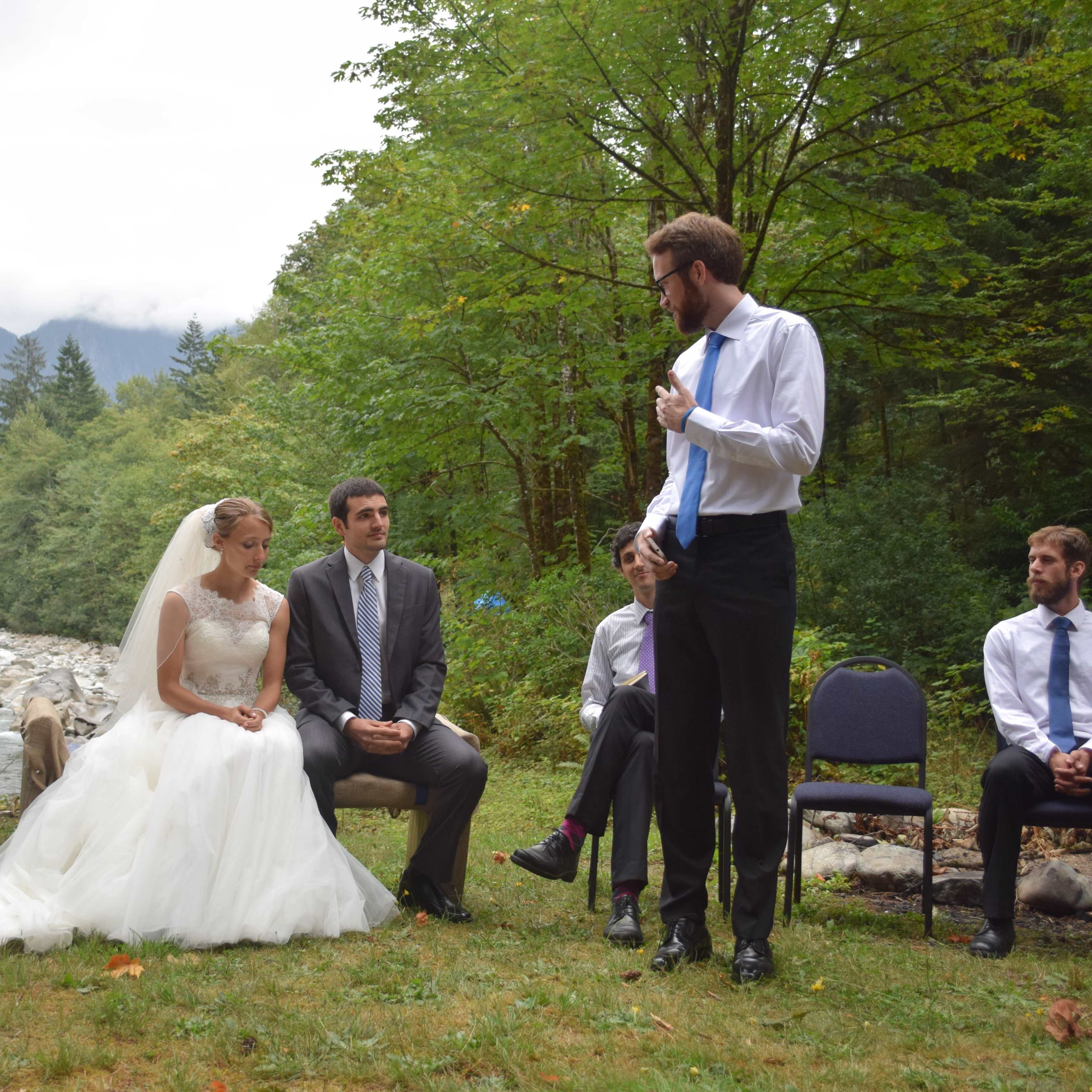 A bride and groom sit on chairs near a river, while a man stands and speaks in front of them, with guests seated nearby in a forested area.