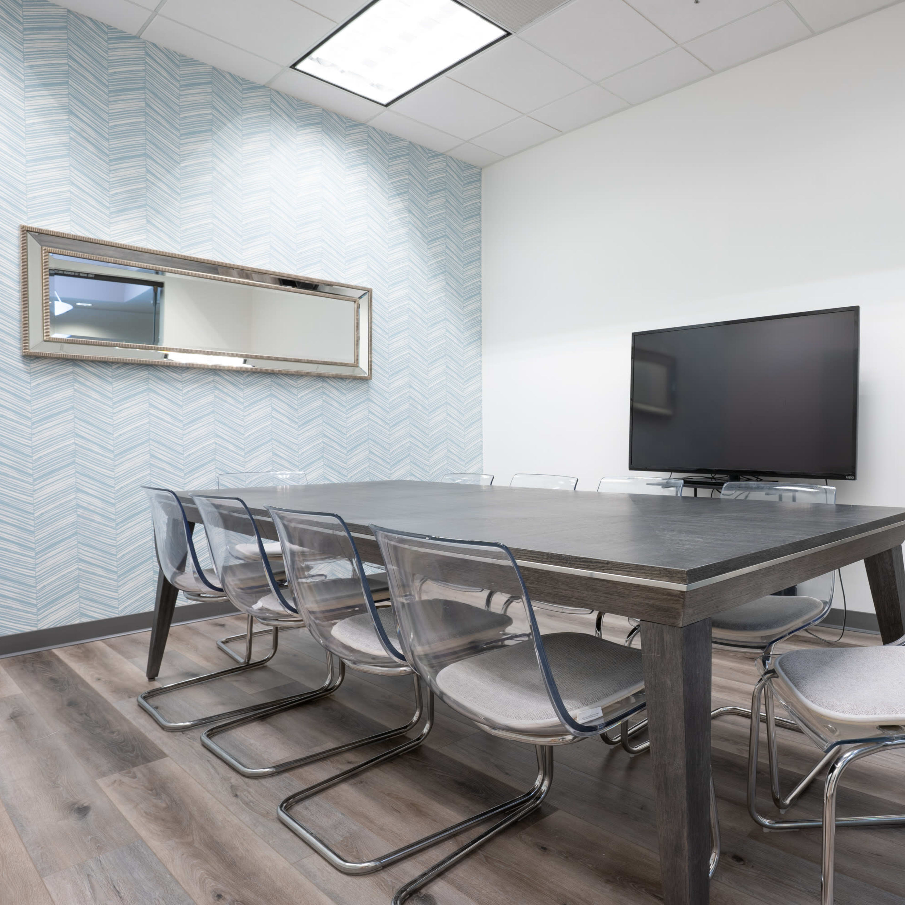 A conference room with a long wooden table surrounded by transparent chairs, a large wall-mounted television, and a coffee station on a side table.