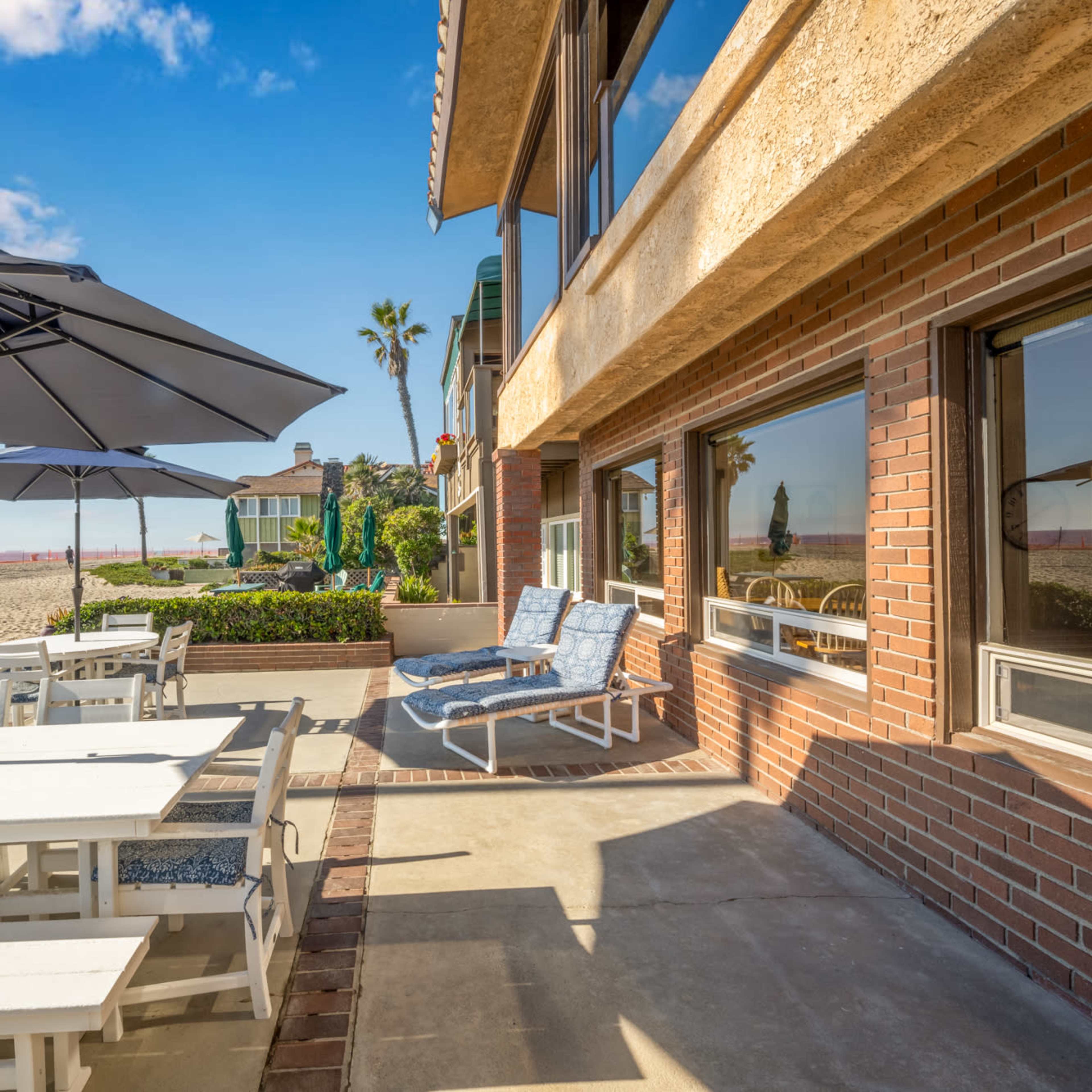 A bright living space features woven furniture, a round wooden table, and large windows showcasing a beach view with umbrellas outside.