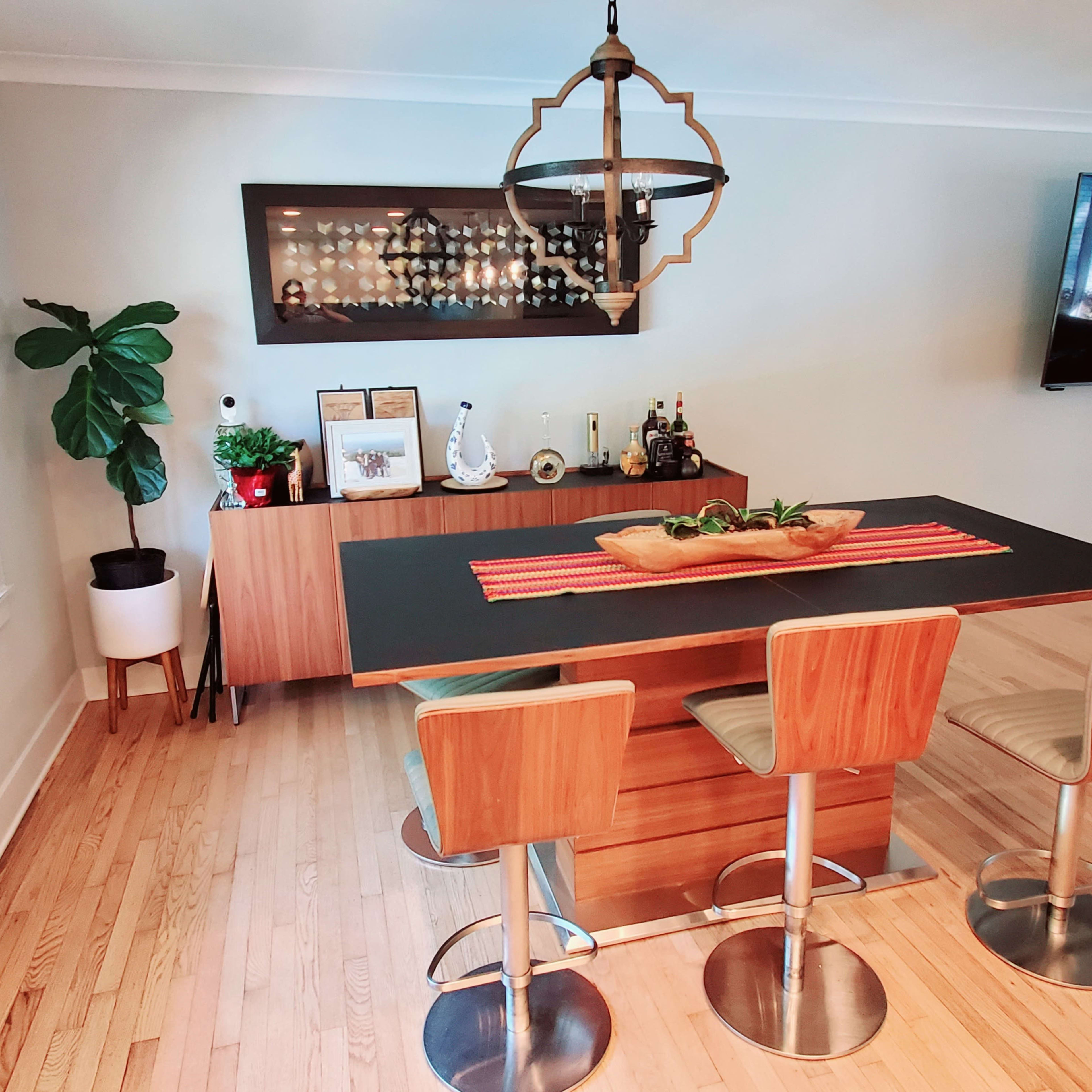 A modern dining area features a rectangular table with three stools, a wall-mounted display case, and a television in the background.