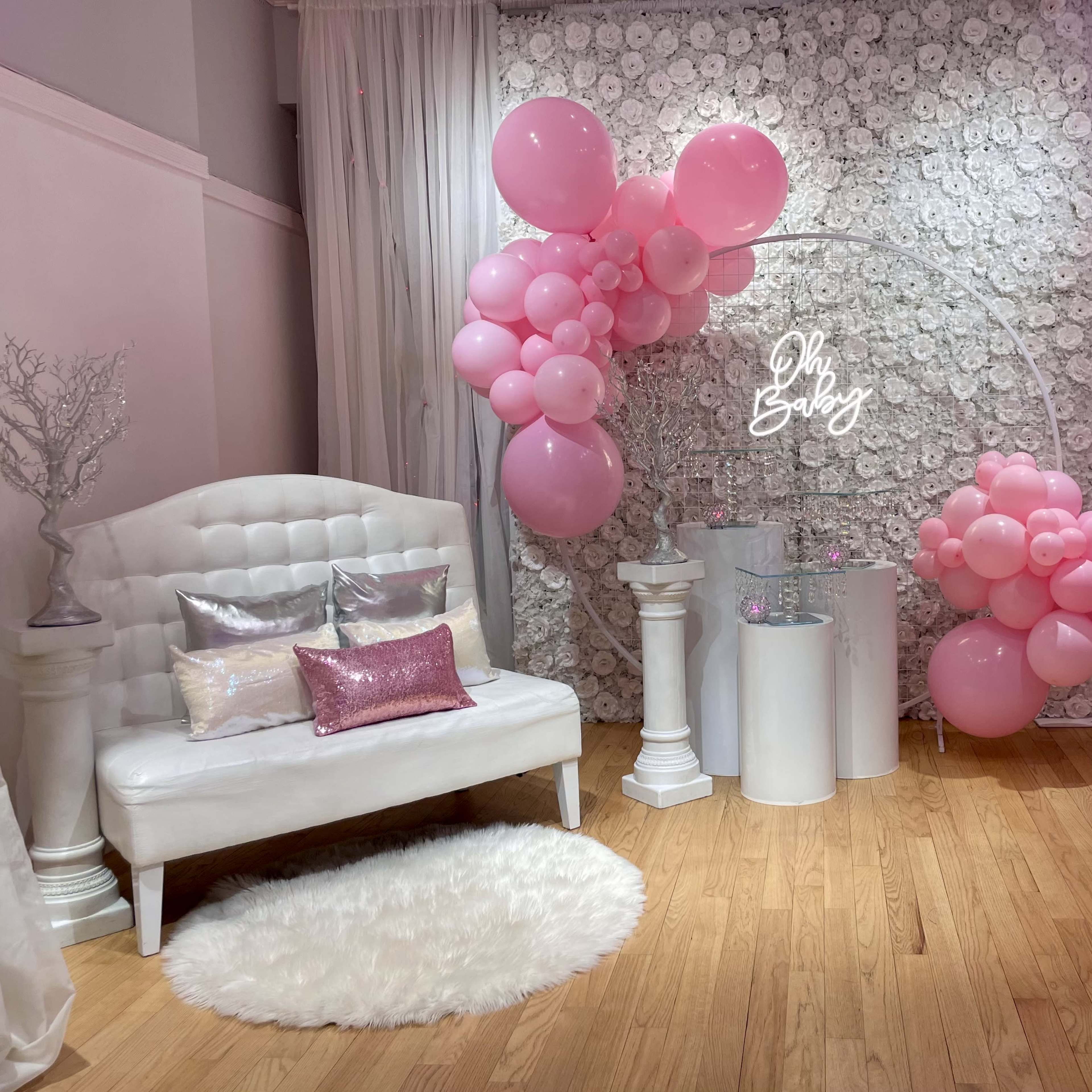 A decorated space for a baby shower, featuring a white couch, a floral backdrop, and pink balloons arranged in an arch.