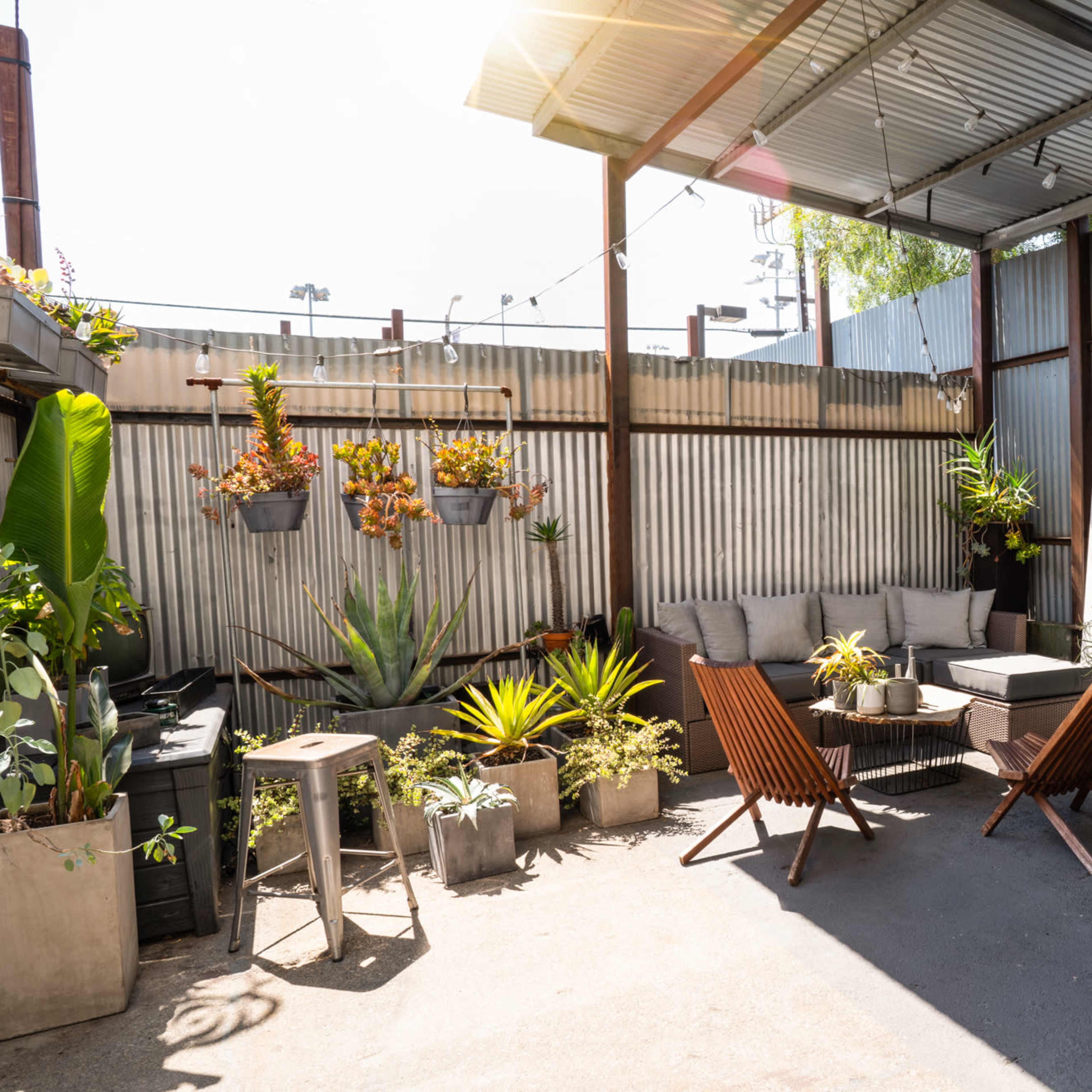 A small, outdoor seating area with two wooden lounge chairs, a sectional sofa, potted plants, and string lights under a metal canopy.