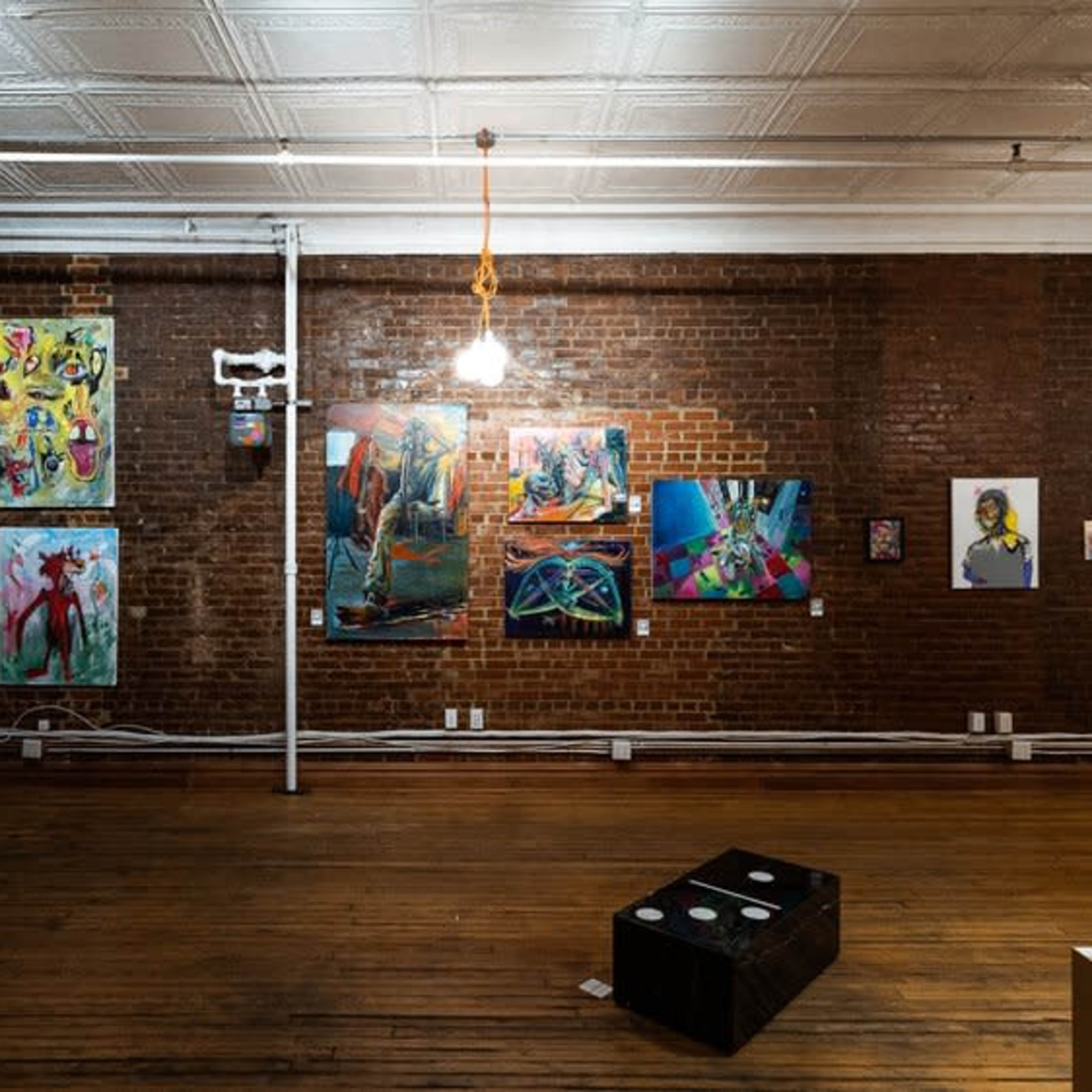 An art gallery with exposed brick walls displaying colorful paintings and sculptures, illuminated by pendant lights.