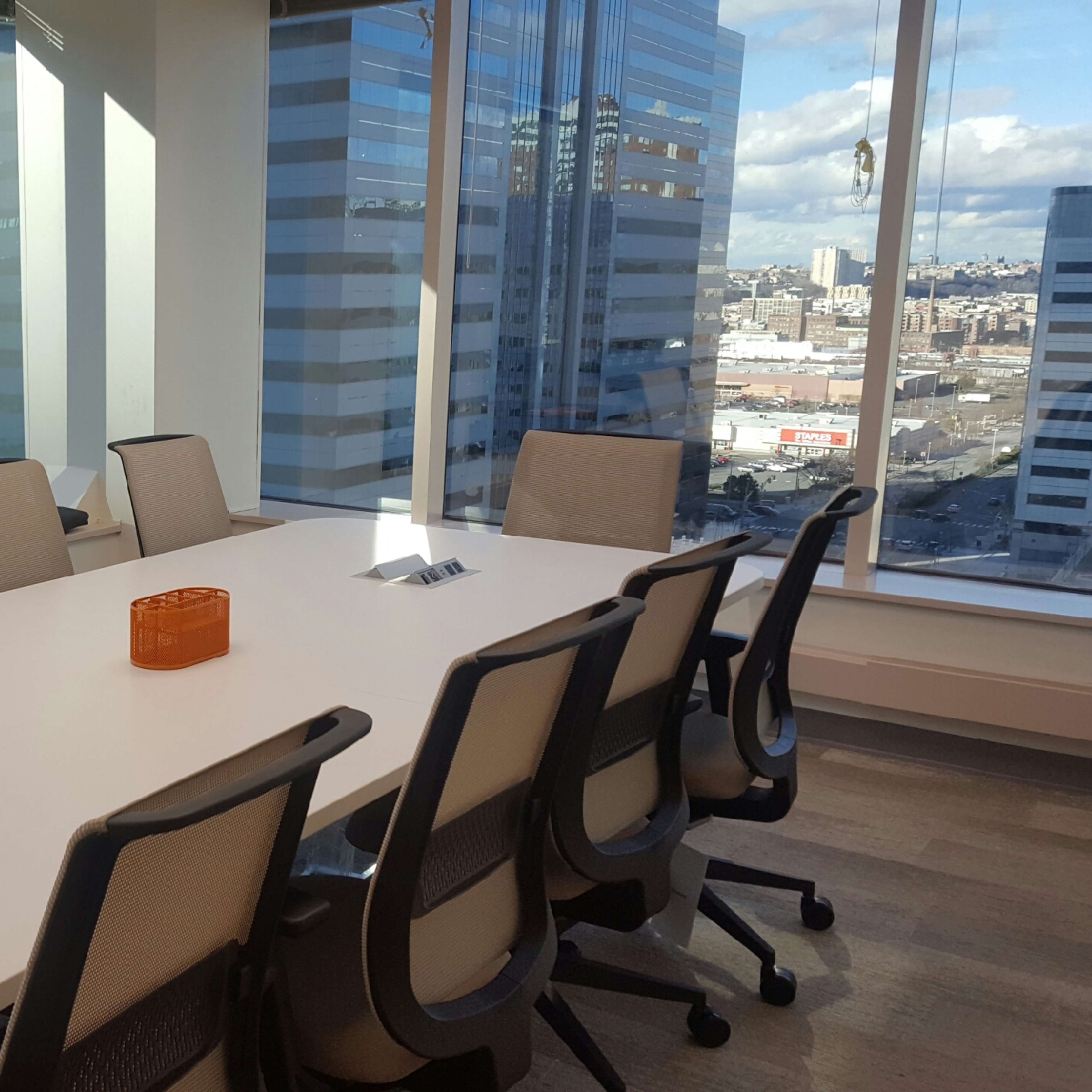 A large conference room features a white table surrounded by office chairs, with floor-to-ceiling windows offering a view of a city skyline.