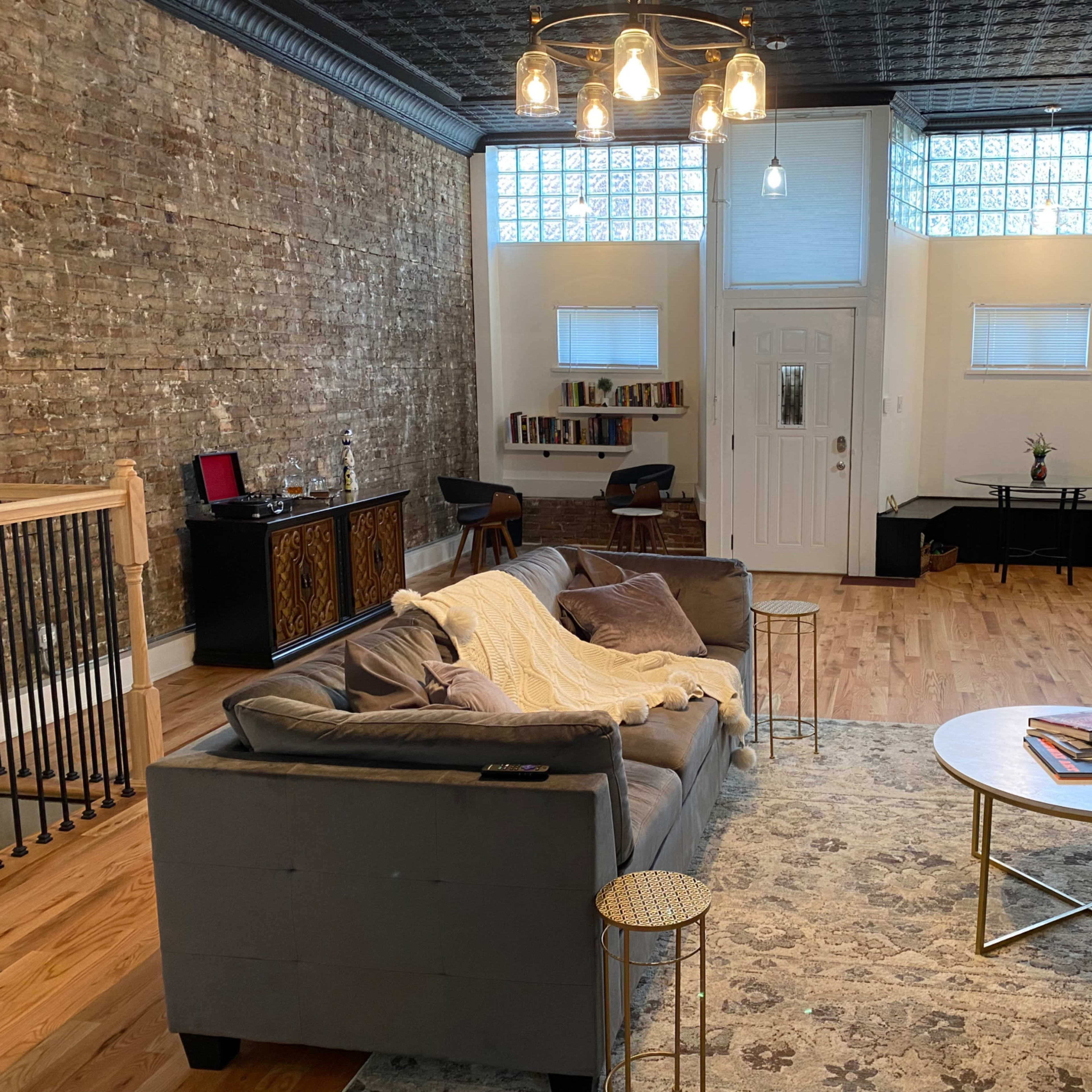 A spacious living room with a brick wall, hardwood flooring, and a sofa facing a round coffee table, illuminated by pendant lights above.