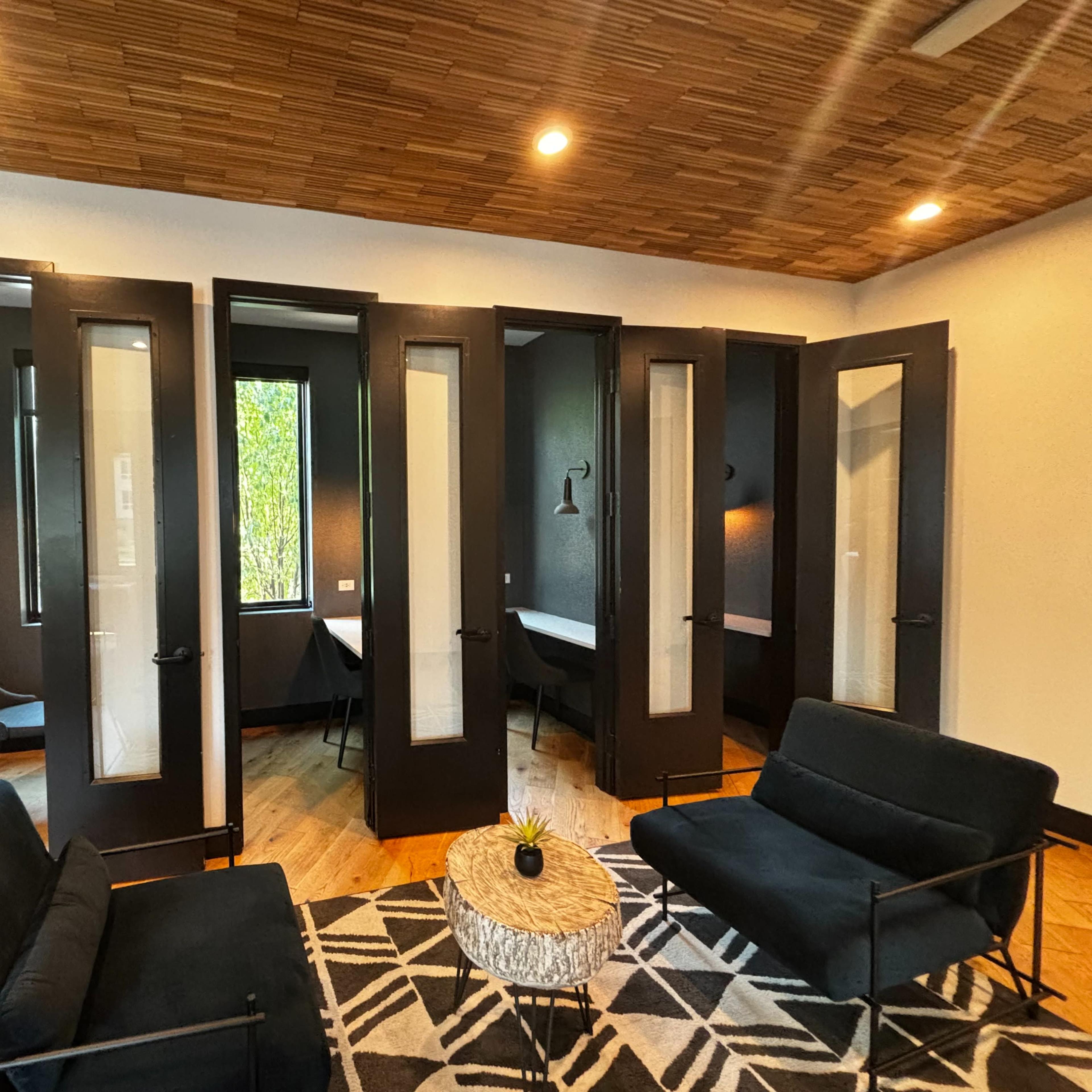 A modern room with dark doors and large windows, featuring two black sofas and a round coffee table on a patterned rug.