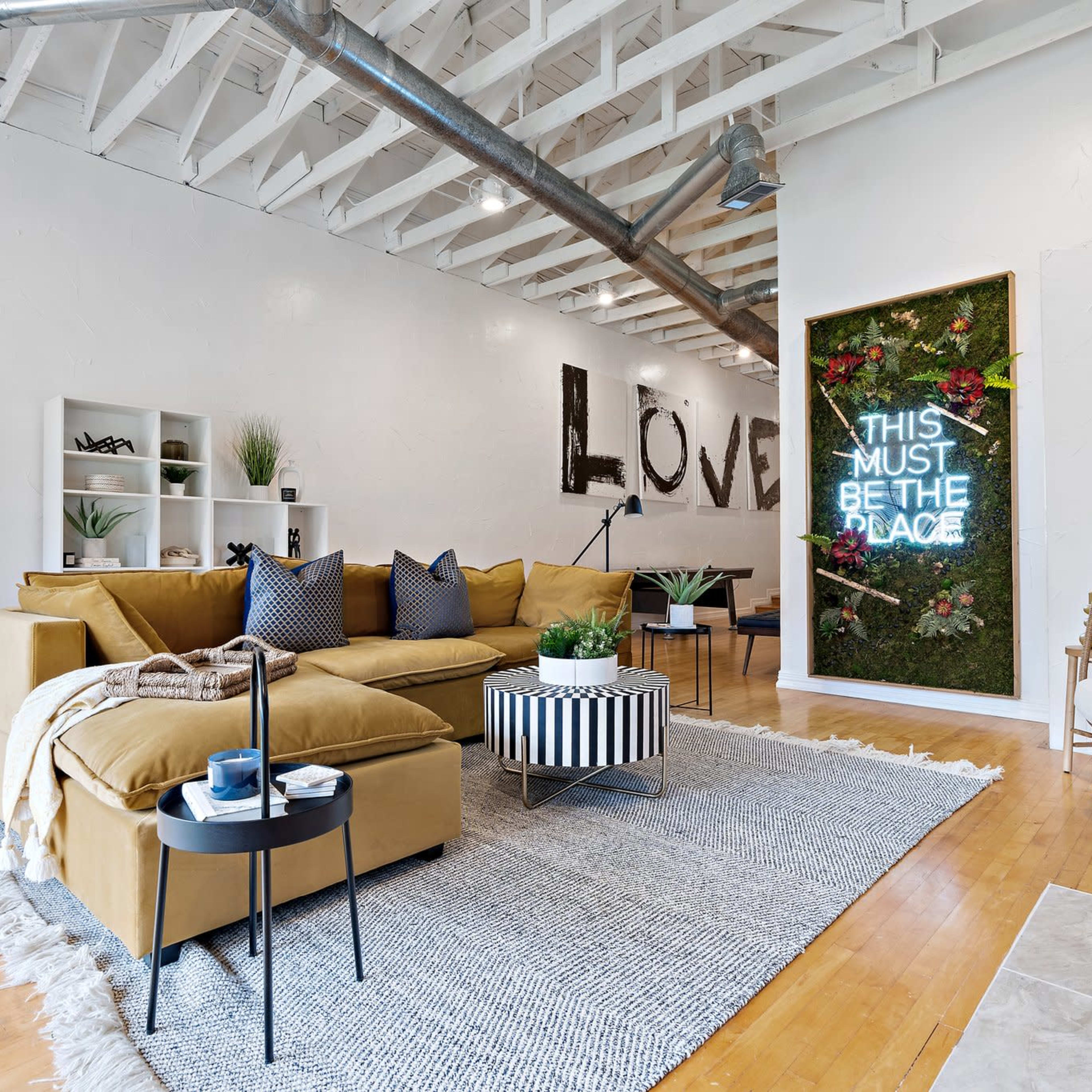 A bright living room features a large yellow sectional sofa, a striped coffee table, and a decorative wall with a neon sign and a "LOVE" mural.