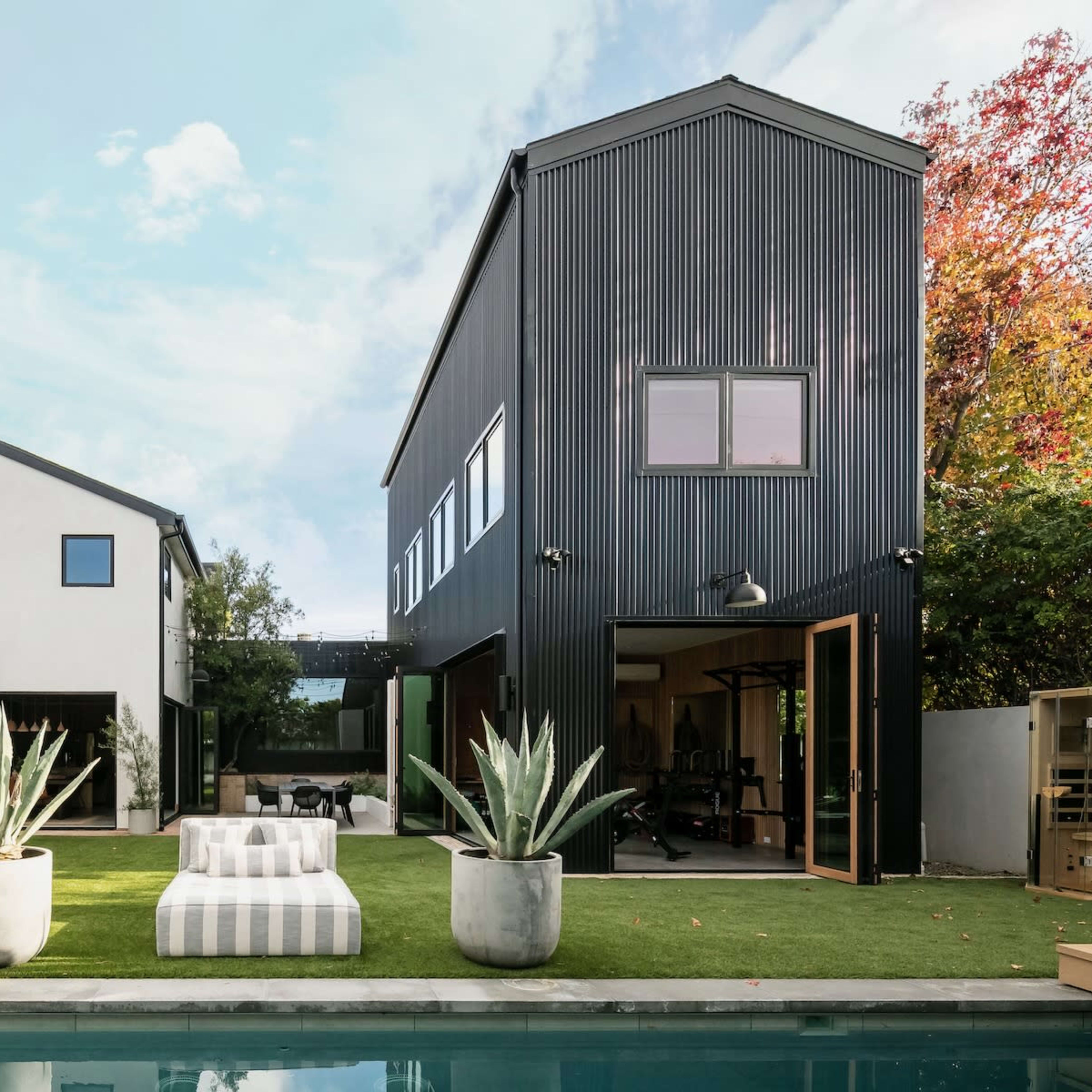 A modern house with a black corrugated metal exterior, accompanied by a white house, a pool, and landscaped seating areas with large planters.
