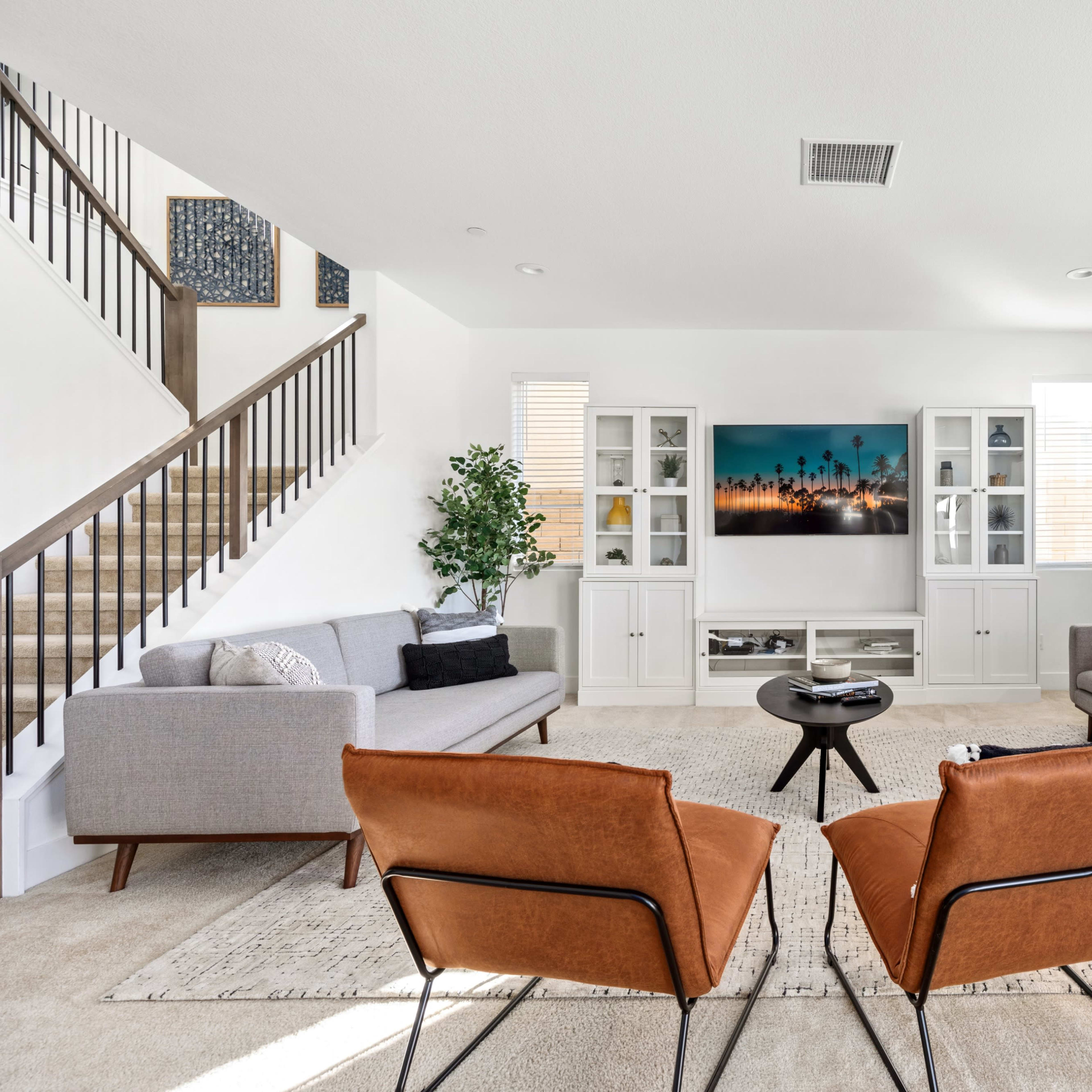 A modern living room features two brown armchairs facing a circular black coffee table, flanked by a gray sofa and a second gray couch, with a television mounted on the wall.