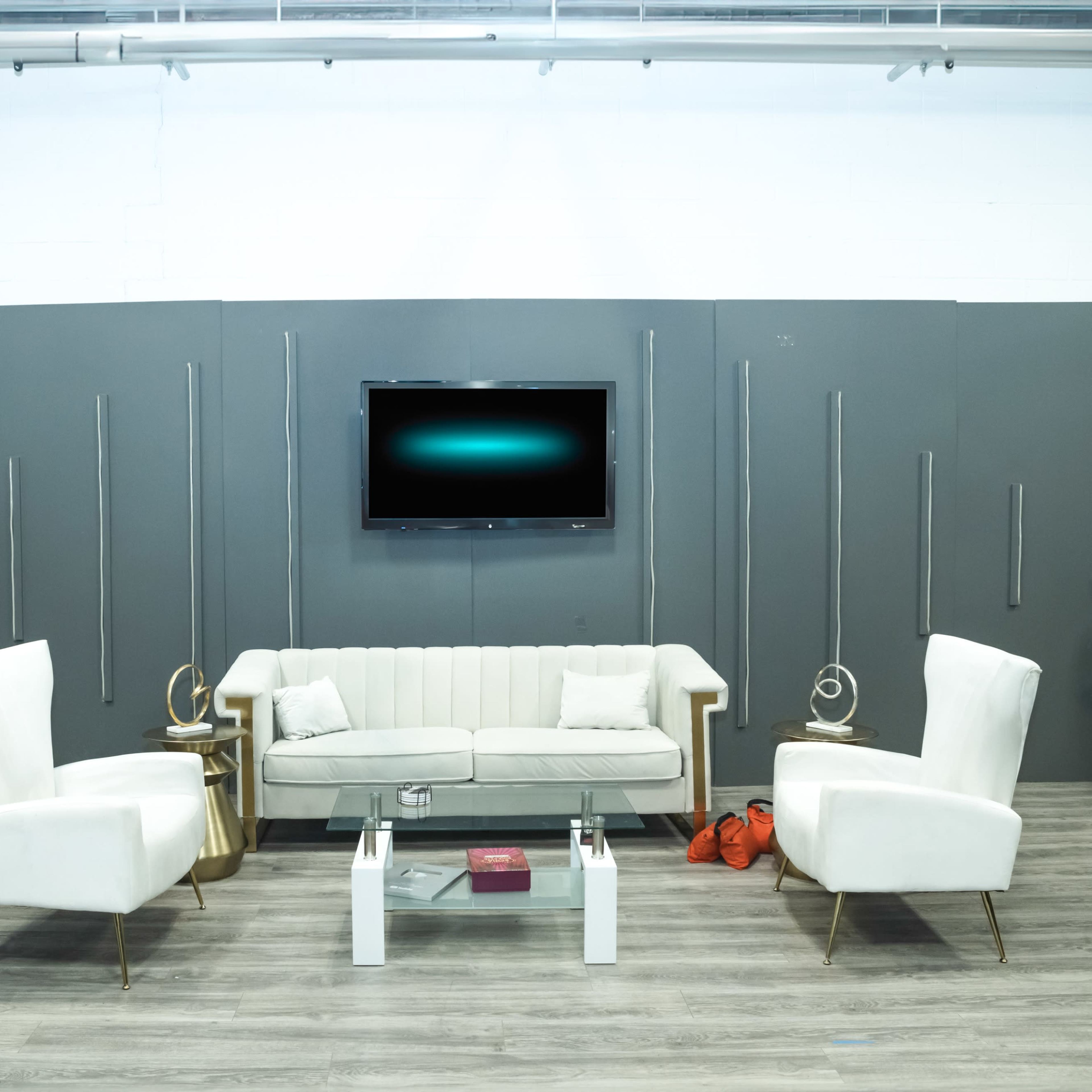 The scene features a modern seating arrangement with a light-colored sofa and two matching armchairs in front of a mounted television, surrounded by a gray wall and a green plant.