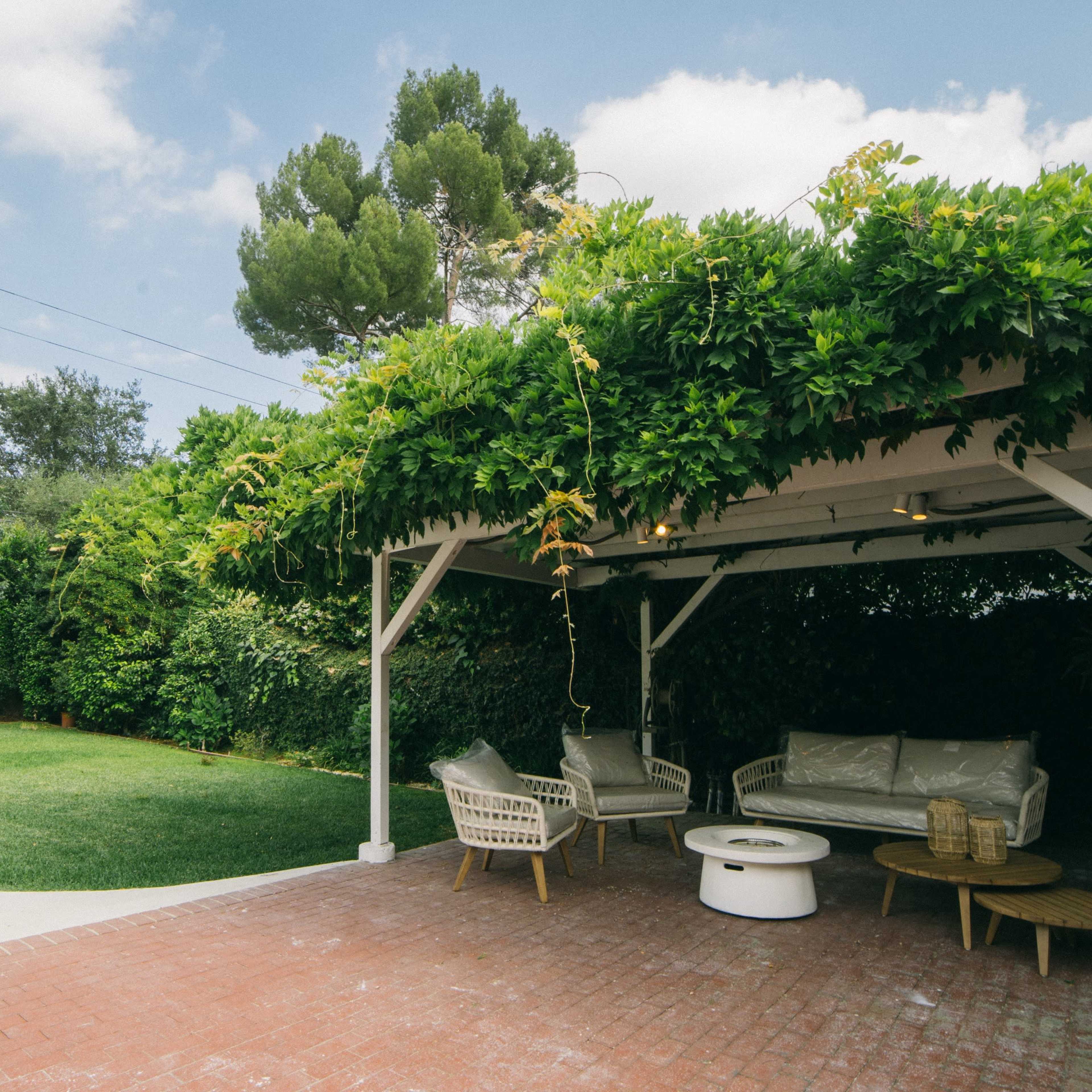 A covered patio with seating and a round table is surrounded by a grassy area and lush greenery under a partly cloudy sky.