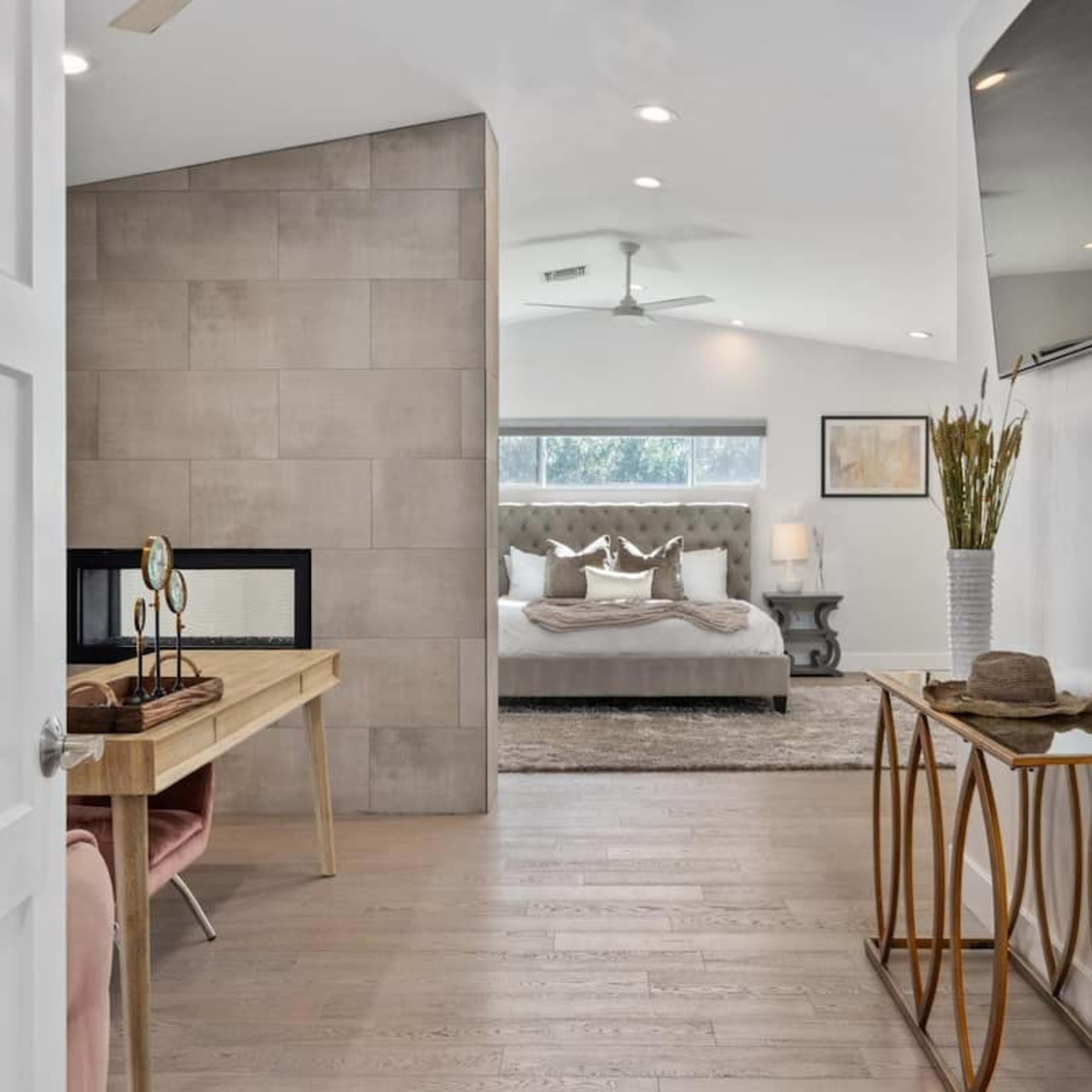 A modern bedroom visible through an open doorway, featuring a bed with decorative pillows, a fireplace, and a minimalist desk with elegant furnishings.