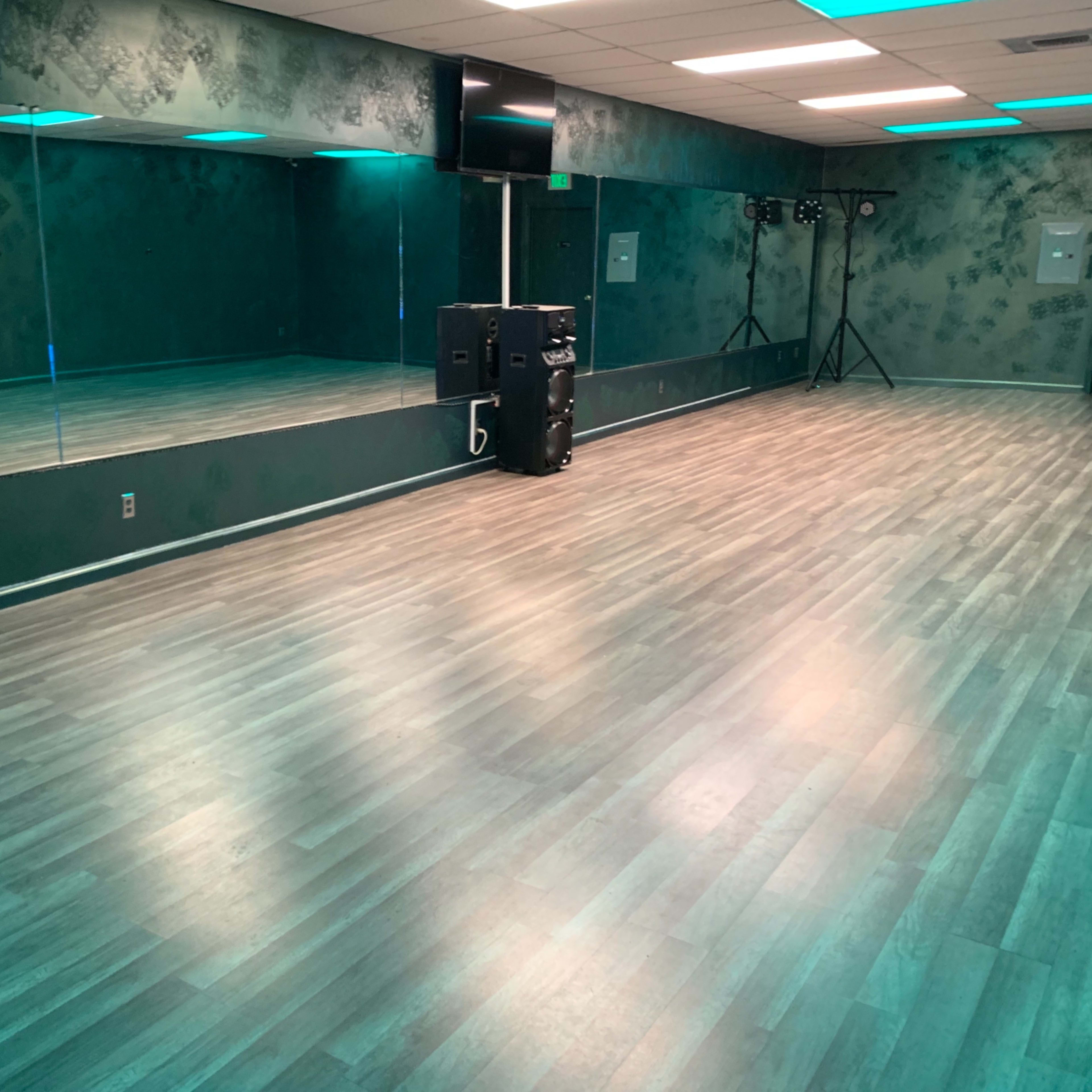 A dance studio with a mirrored wall, wooden flooring, and teal lighting effects.