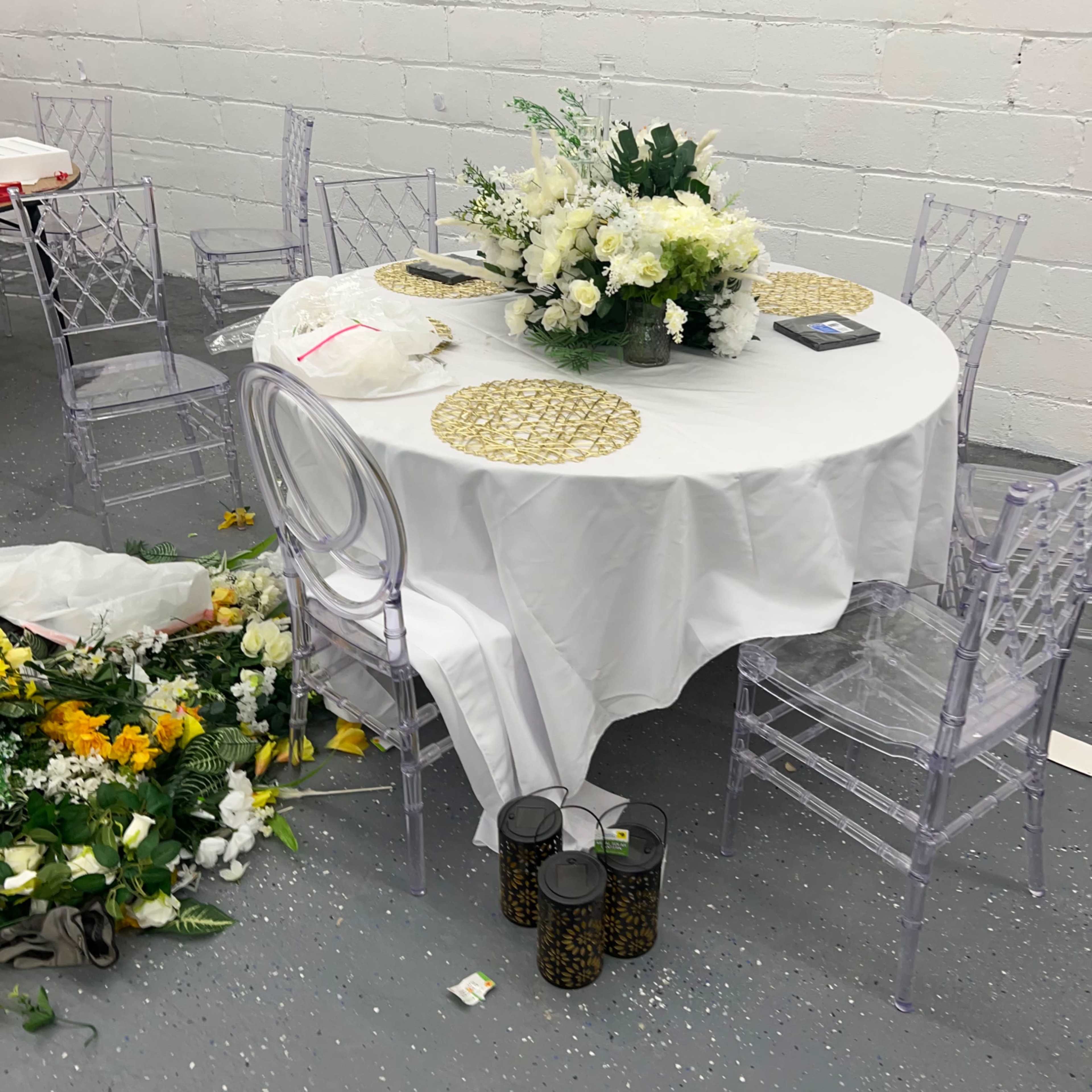 A round table covered with a white tablecloth is set with floral centerpieces and surrounded by clear chairs, while a pile of artificial flowers is scattered on the floor nearby.