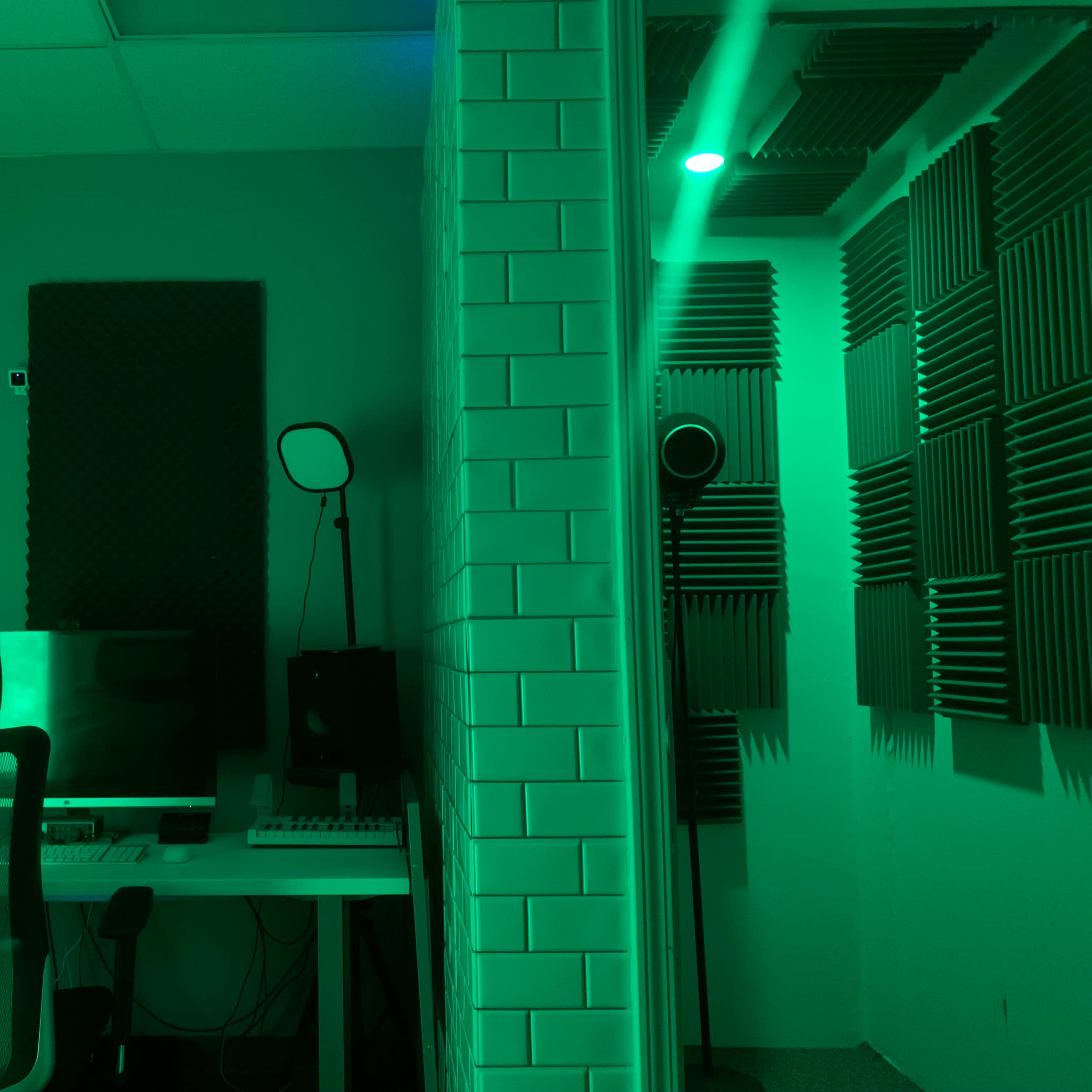 A soundproof recording studio features a desk with a computer and audio equipment, surrounded by acoustic panels and a green ambient light.