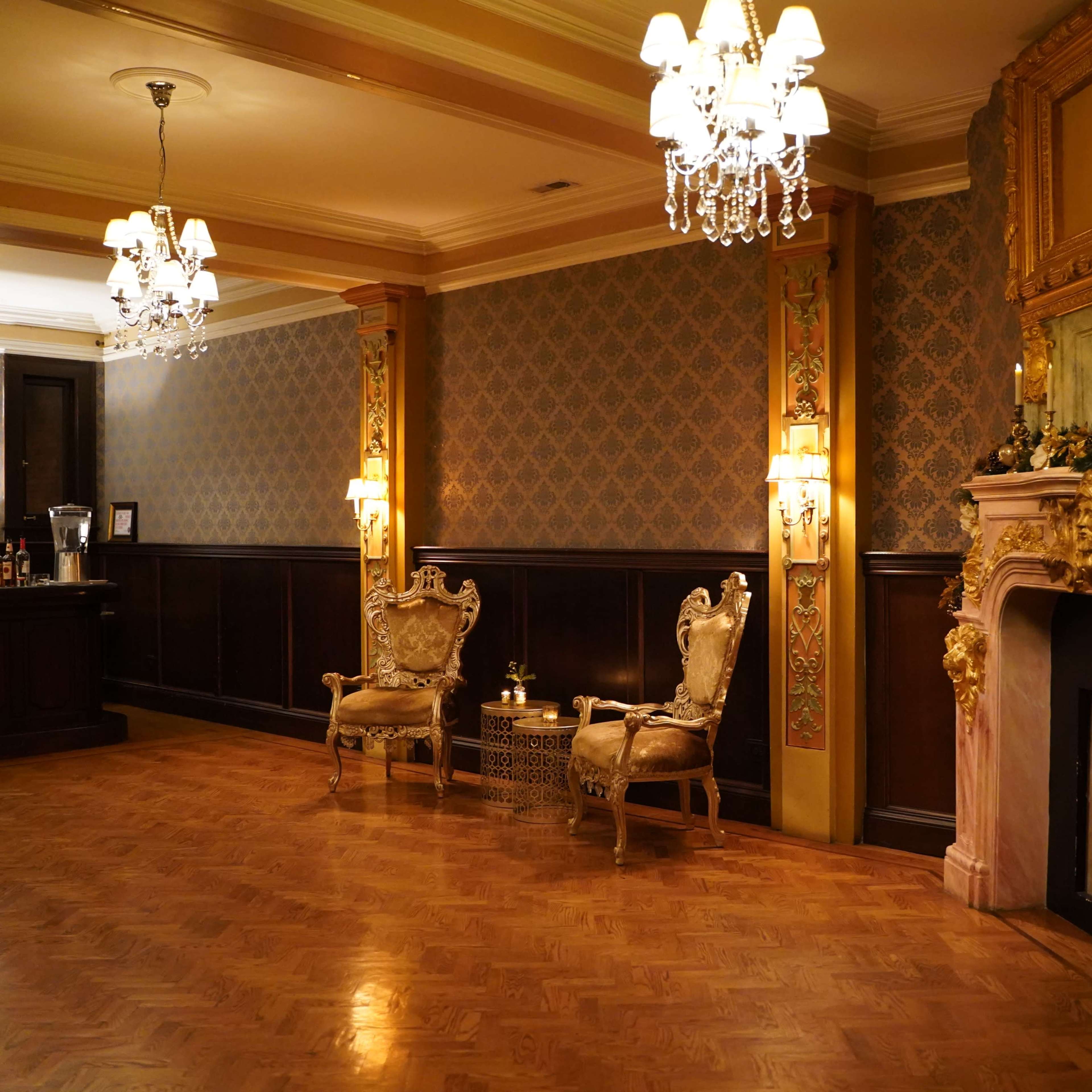 An ornate room with a patterned wallpaper, a carved fireplace adorned with decorative elements, and two elegant chairs placed beside a small table.