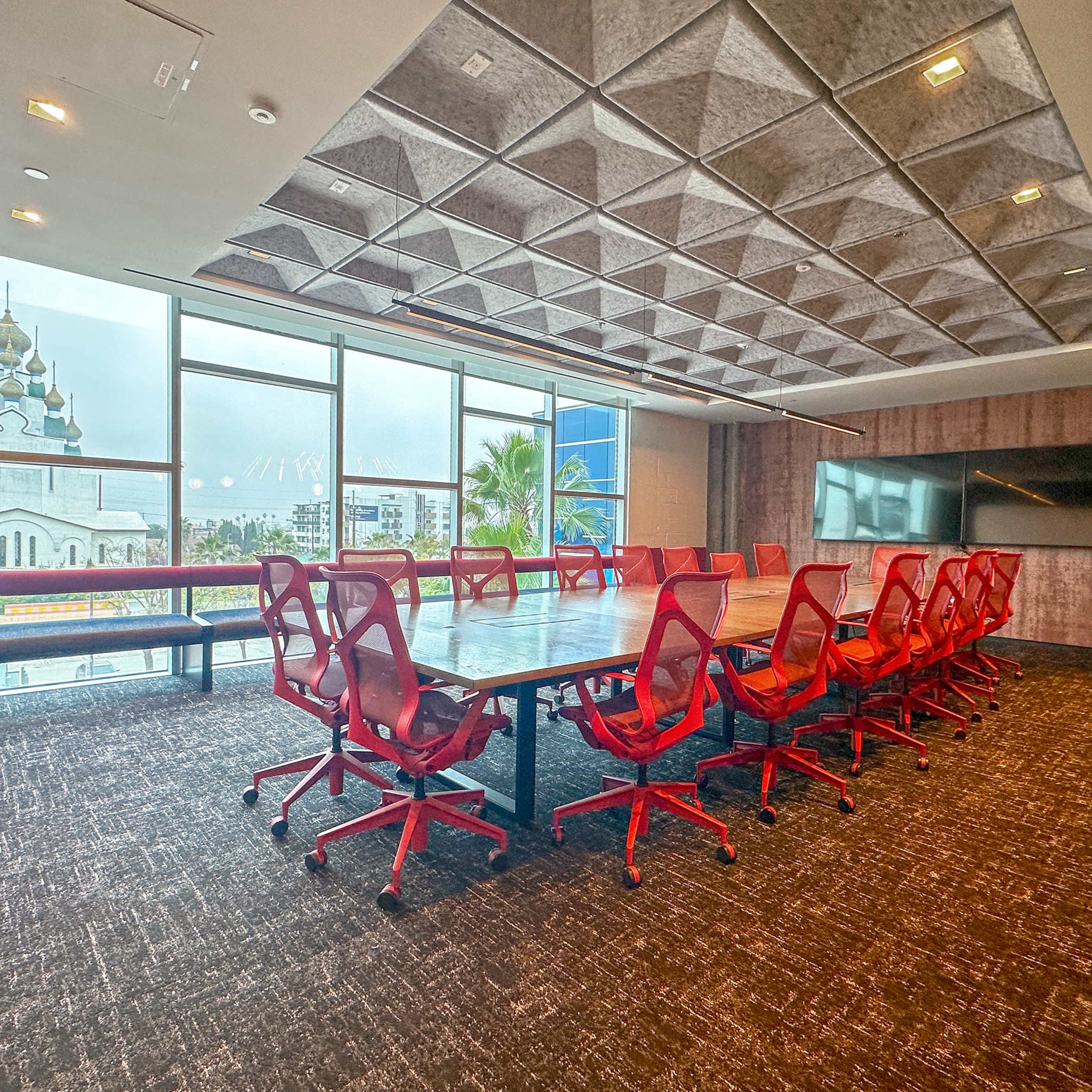 A conference room features a large table surrounded by red chairs, with large windows providing a view of an outdoor area.