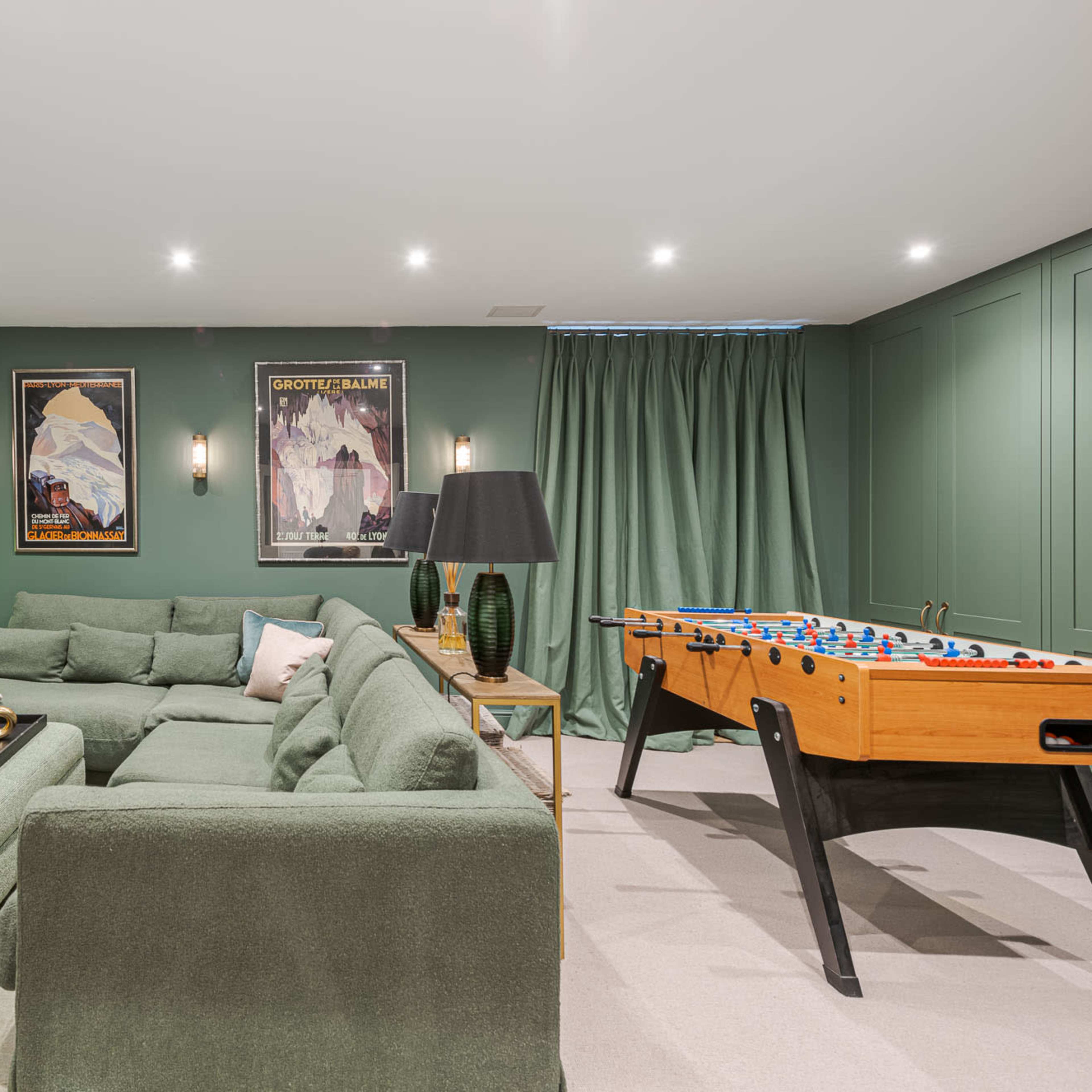 A modern basement lounge area featuring a green sectional sofa, a foosball table, built-in shelves with books, and framed posters on the walls.