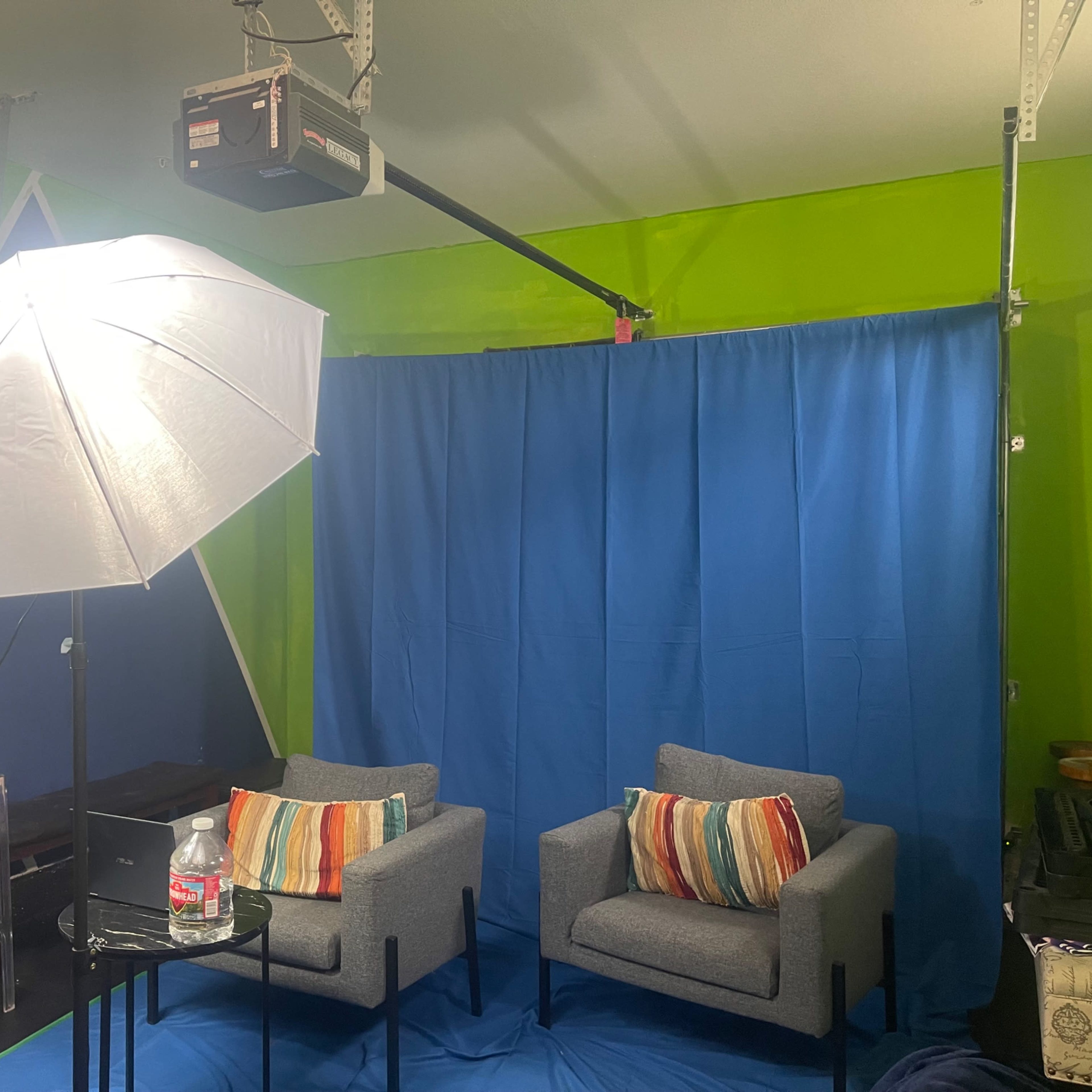The scene features a setup with two gray chairs and a small glass table in front of a blue backdrop, illuminated by soft lighting from an umbrella light.