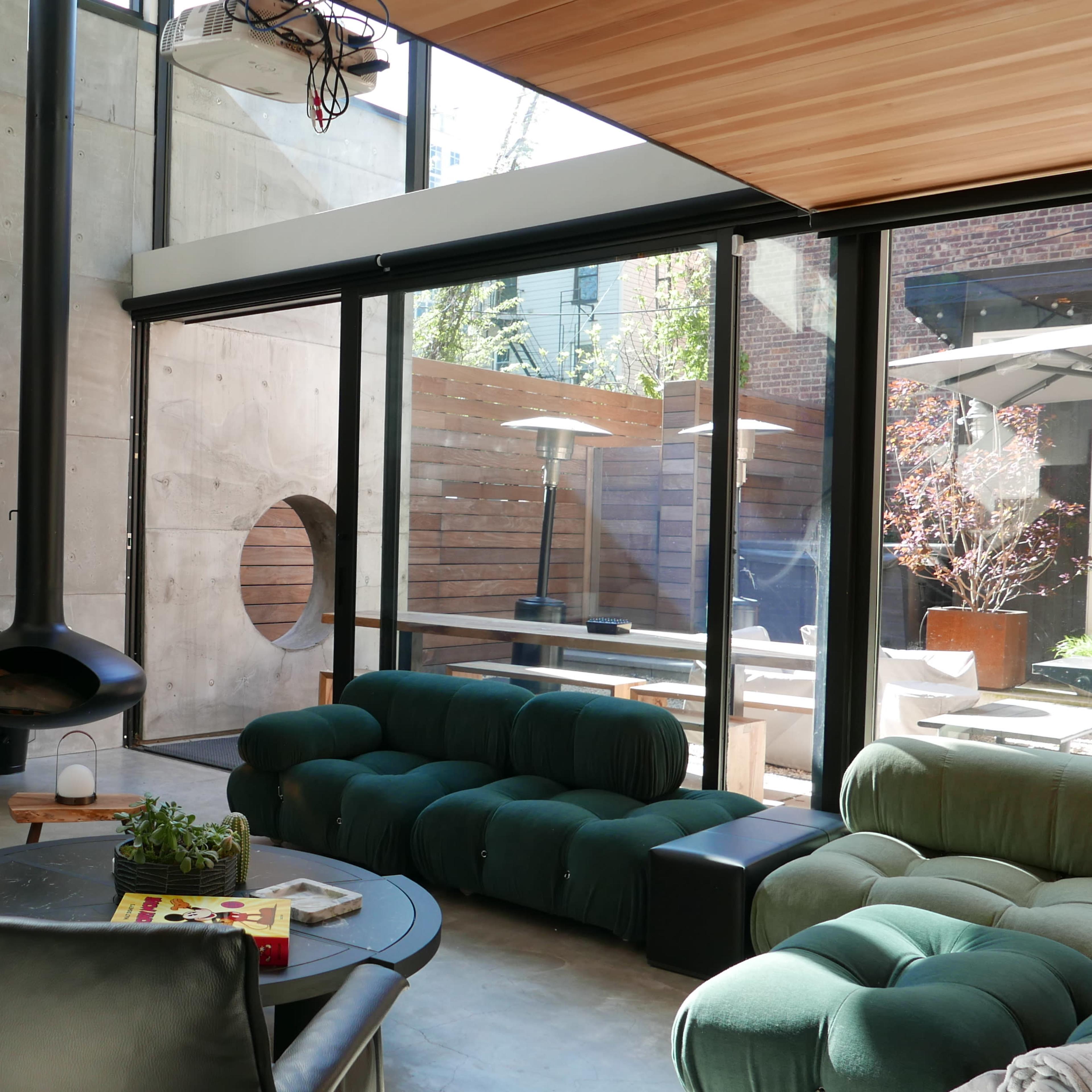 The living room features a modern design with a green sofa, a black TV stand, a circular fireplace, and large windows opening to an outdoor patio.