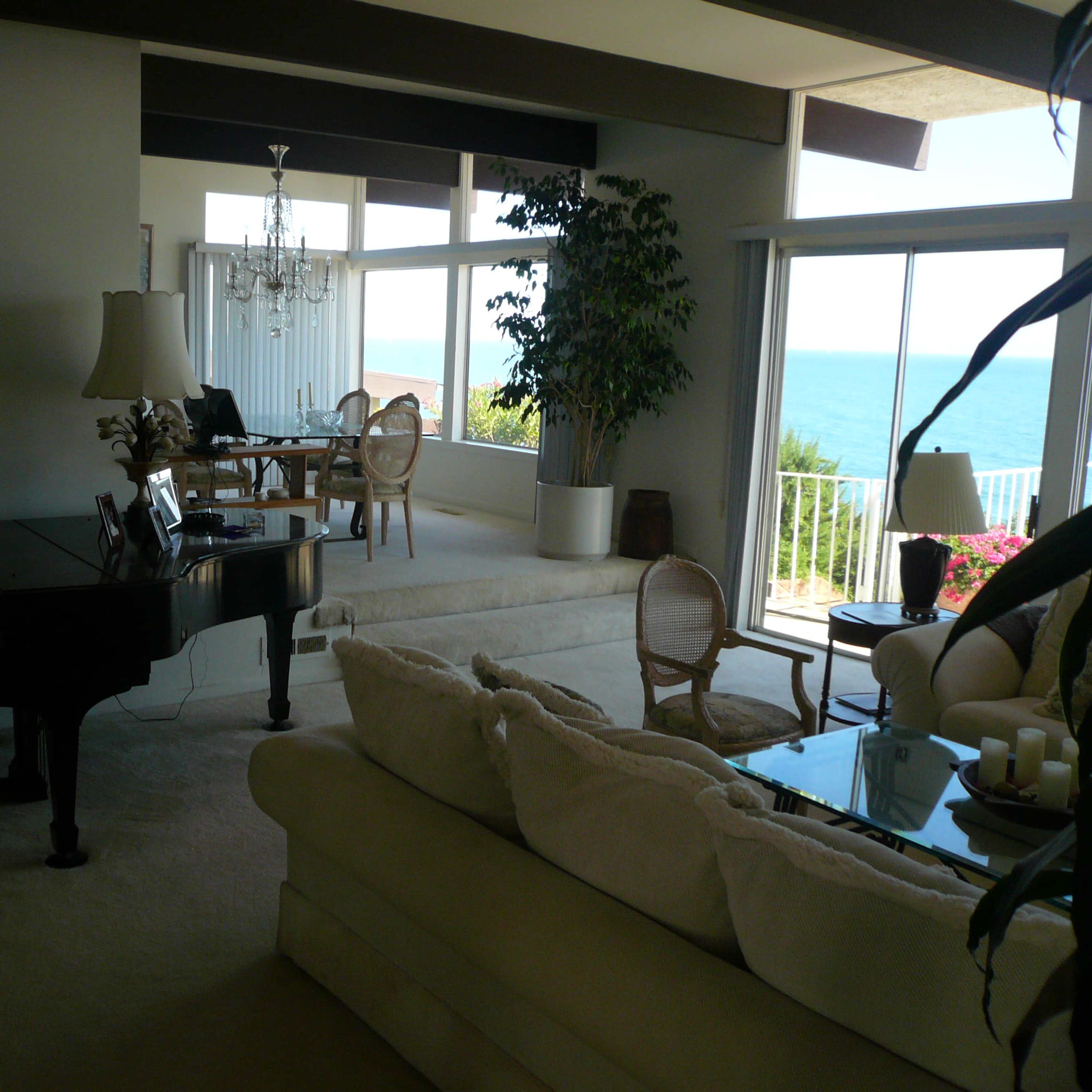 A spacious living room with a large window overlooking the ocean, featuring a grand piano, a comfortable sofa, and a dining area in the background.